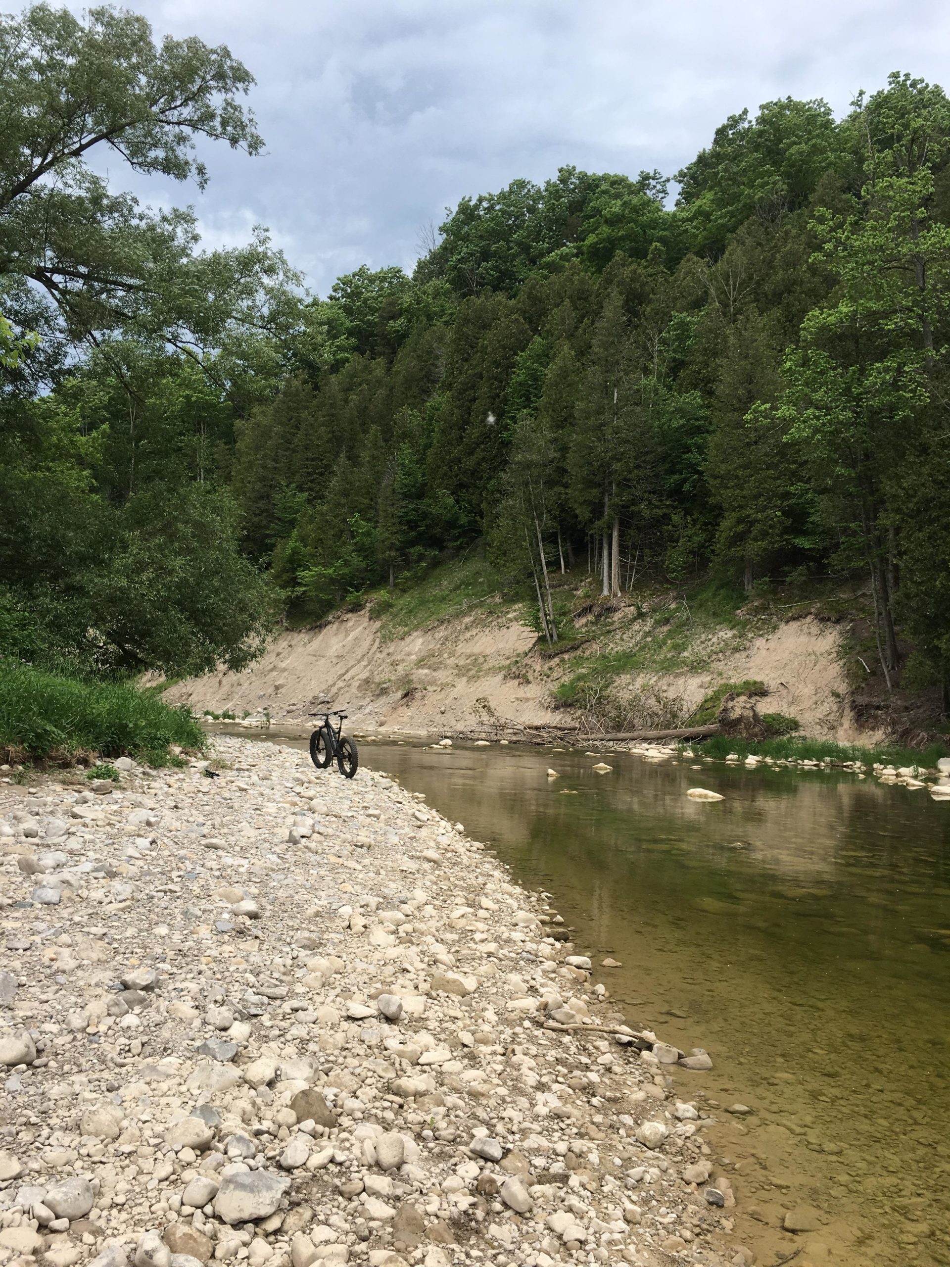 A tranquil riverside scene featuring a gravelly bank with scattered stones, a calm waterway reflecting the surrounding greenery, and a bicycle resting beside the shore. Tall trees and a hillside dense with foliage rise in the background under a cloudy sky. Bayfield river mountain bike trail.