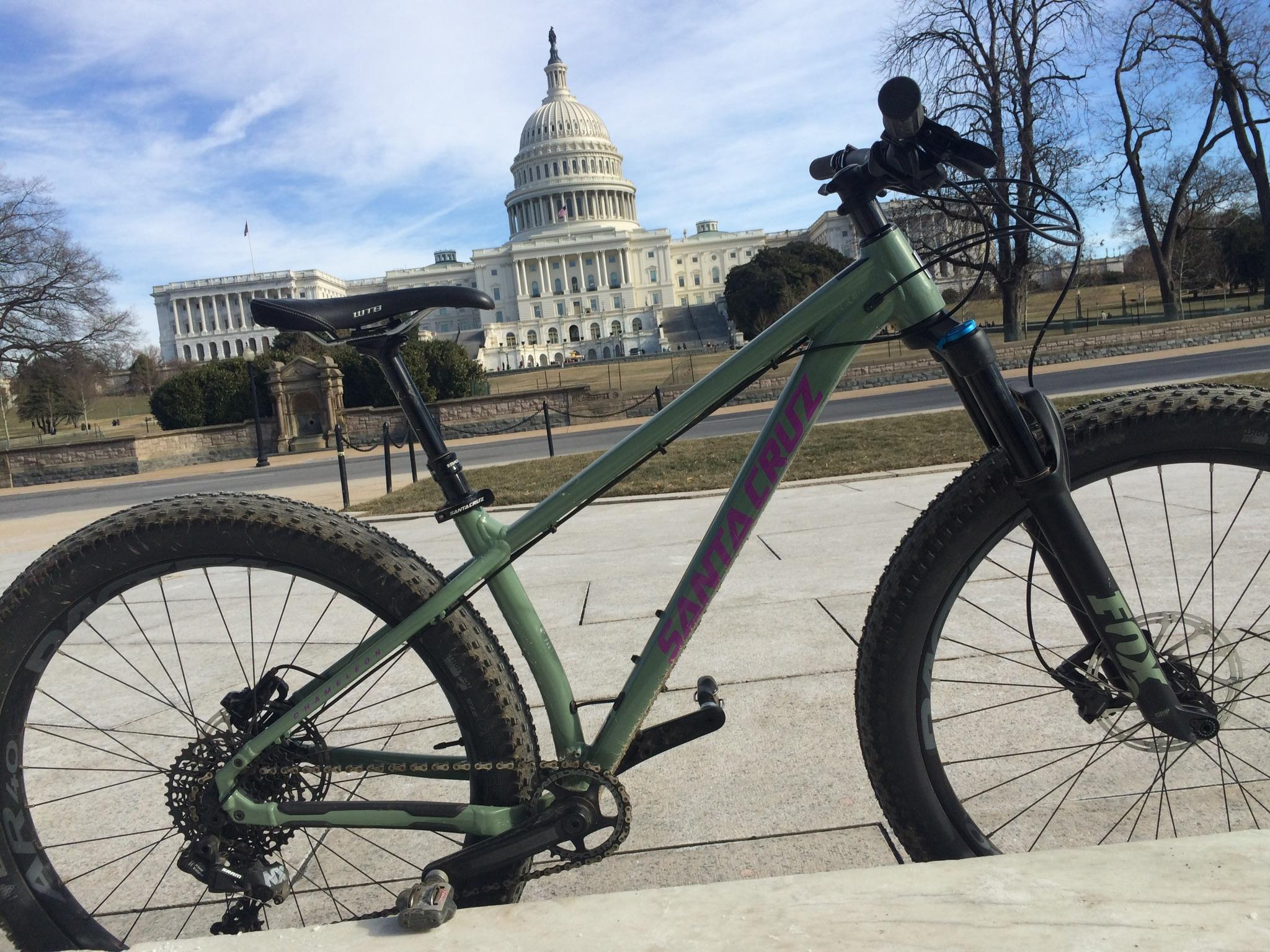 Santa Cruz Chameleon: A Santa Cruz mountain bike is positioned in the foreground, with the United States Capitol building in the background. The bike features a green frame and is resting on a marble surface, showcasing its large tires and detailed components. The sky is partly cloudy, and the surrounding area includes grassy lawns and trees.