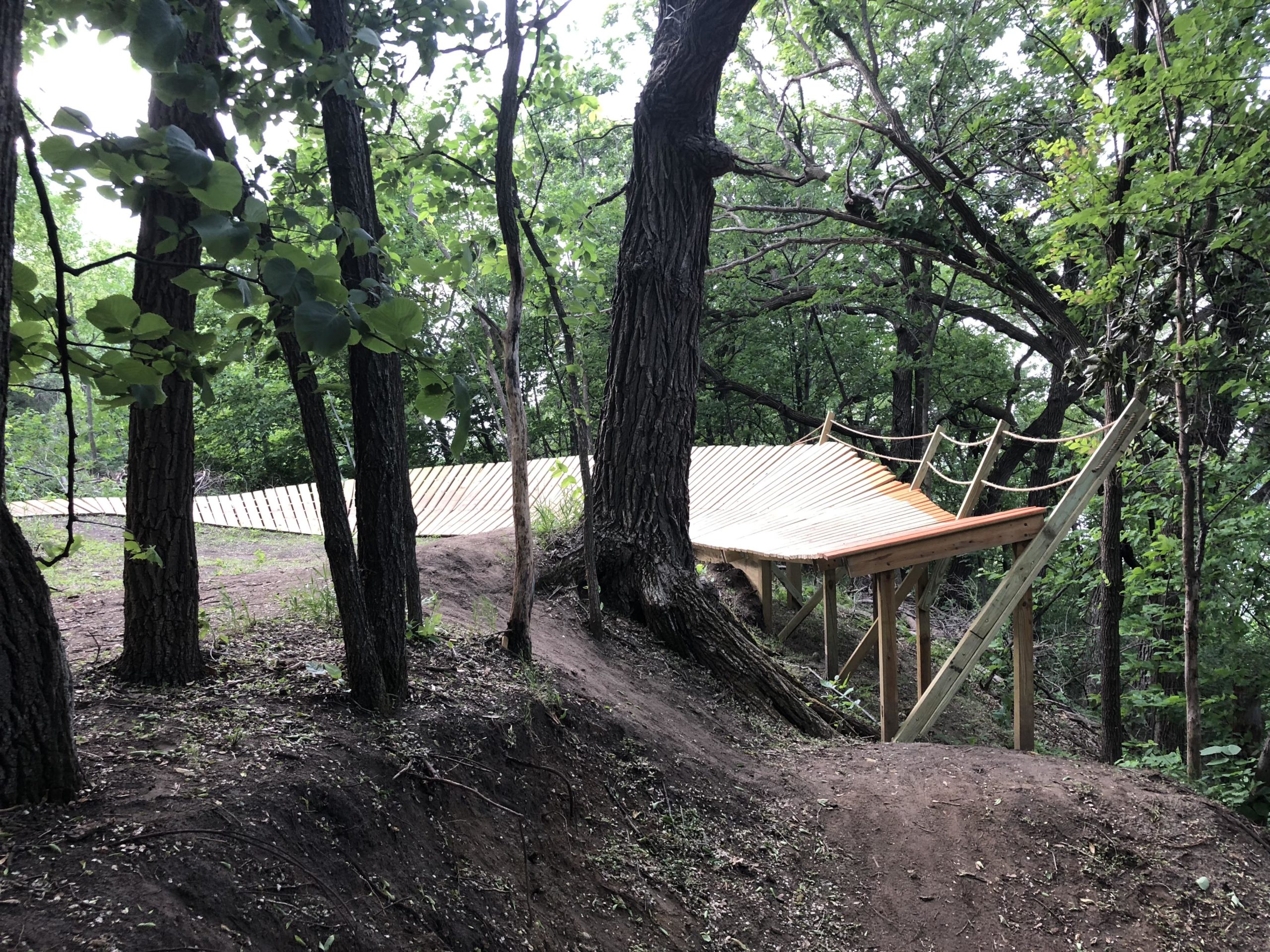 A wooden deck or platform elevated among trees, surrounded by lush green foliage, with a gentle slope leading down to the forest floor. The structure features a sloped roof and is supported by wooden beams, blending into the natural environment. Bertram Chain of Lakes Trail mountain bike trail.