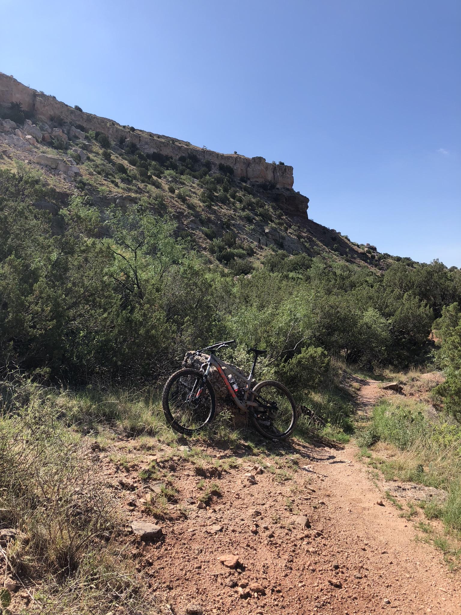 A mountain bike is leaning against a rock on a dirt trail surrounded by green shrubs and hills, with a clear blue sky in the background. Palo Duro Canyon mountain bike trail.