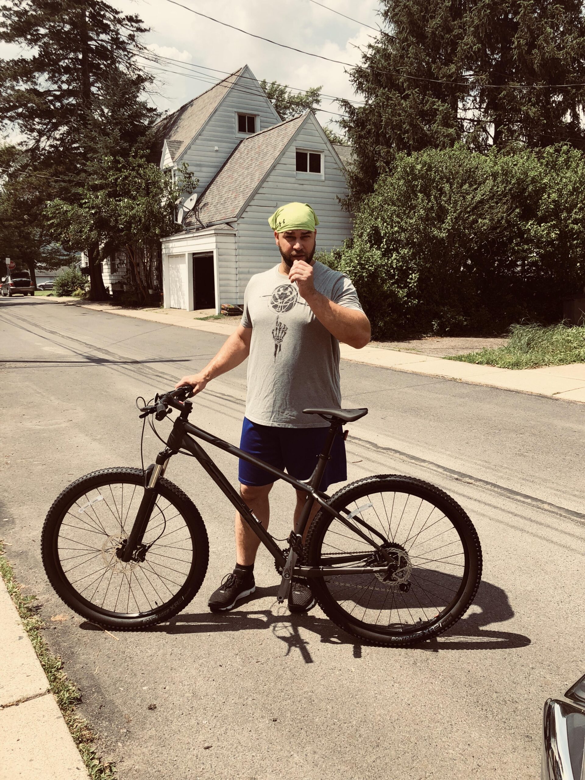 Trek Xcaliber 7: A man wearing a gray t-shirt, blue shorts, and a green bandana stands on a residential street beside a black mountain bike. He is holding a snack in one hand and appears to be chewing while looking towards the camera. The background features a house with a sloping roof and some greenery.