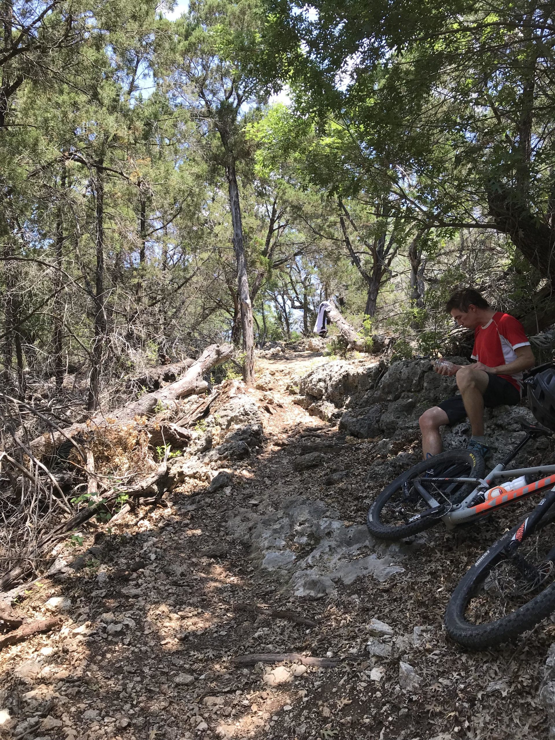 A mountain biker resting on a rocky trail surrounded by dense trees and foliage. Two mountain bikes are parked nearby on the ground, with sunlight filtering through the leaves overhead. Goodwater Trail mountain bike trail.