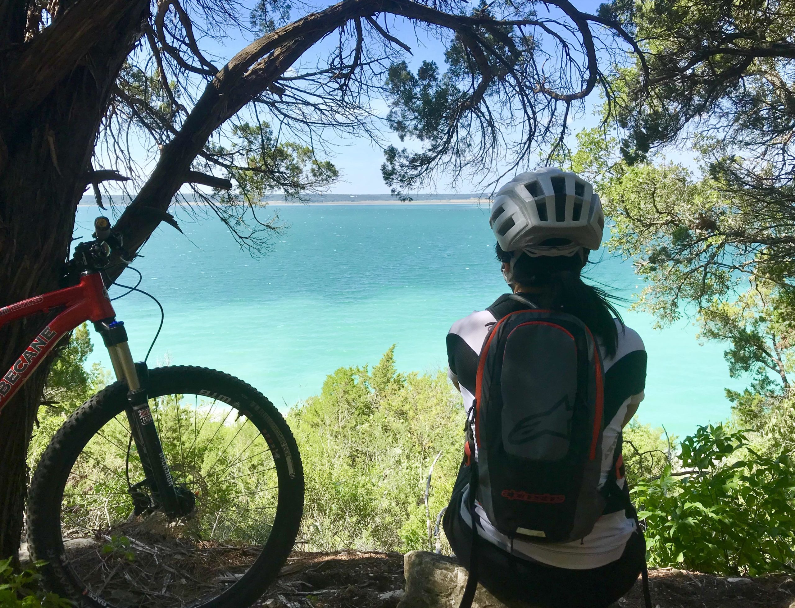 A person sitting on a rock beneath a tree, wearing a helmet and a backpack, gazes out at a tranquil blue-green lake. A mountain bike is resting nearby, surrounded by lush greenery under a clear sky. Dana Peak mountain bike trail.