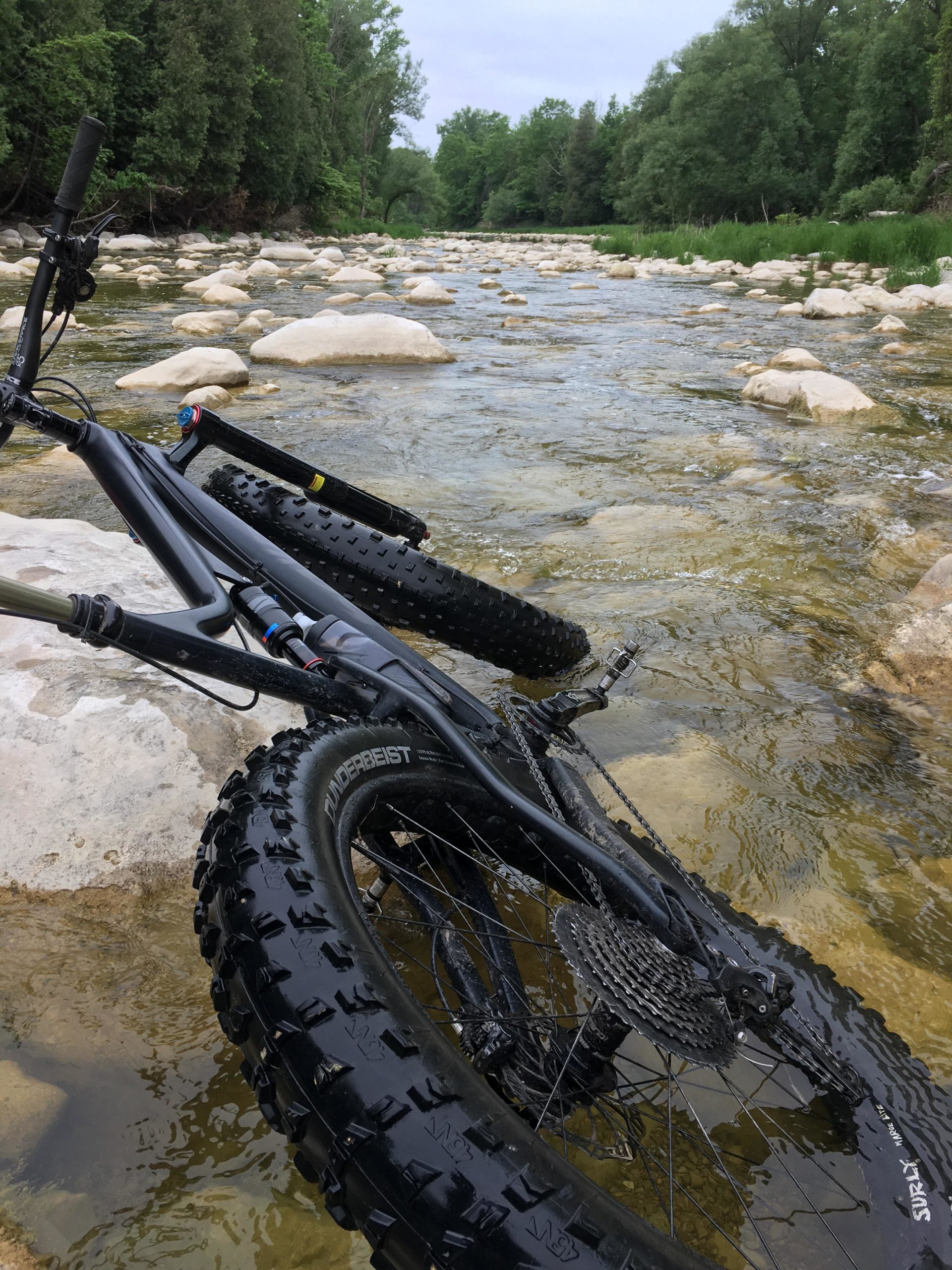 A black mountain bike rests partially submerged in a shallow river, surrounded by smooth, large rocks. The scene is set in a lush, green landscape with trees lining the riverbank, under a cloudy sky. The bike's tires are thick and rugged, designed for off-road terrain. Bayfield river mountain bike trail.