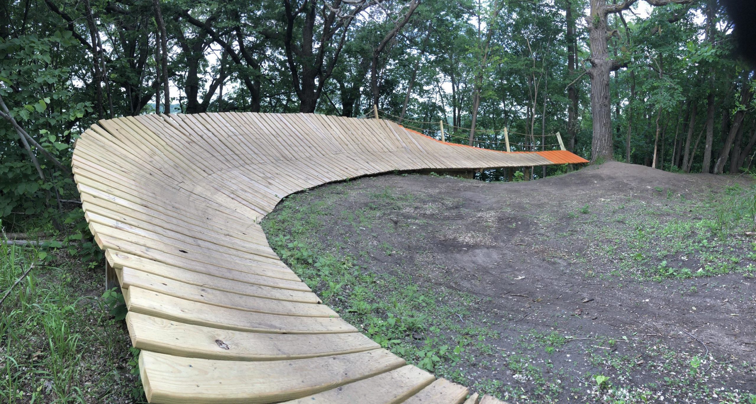 A wooden bike ramp curved along the ground, surrounded by trees and greenery. The ramp features a smooth, spiral design leading up to a small dirt mound. Bertram Chain of Lakes Trail mountain bike trail.