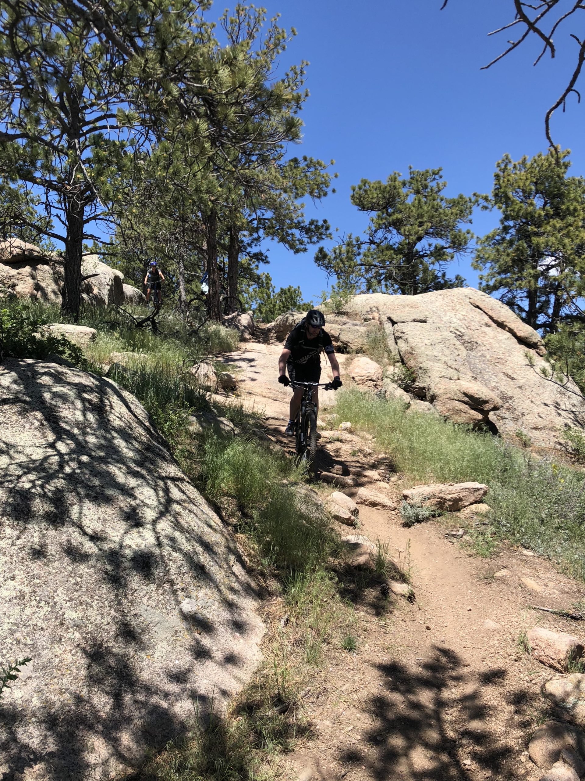 Mountain bikers navigating a rocky trail surrounded by tall trees and clear blue skies. One biker is riding down the path while another is visible in the background. Curt Gowdy State Park mountain bike trail.