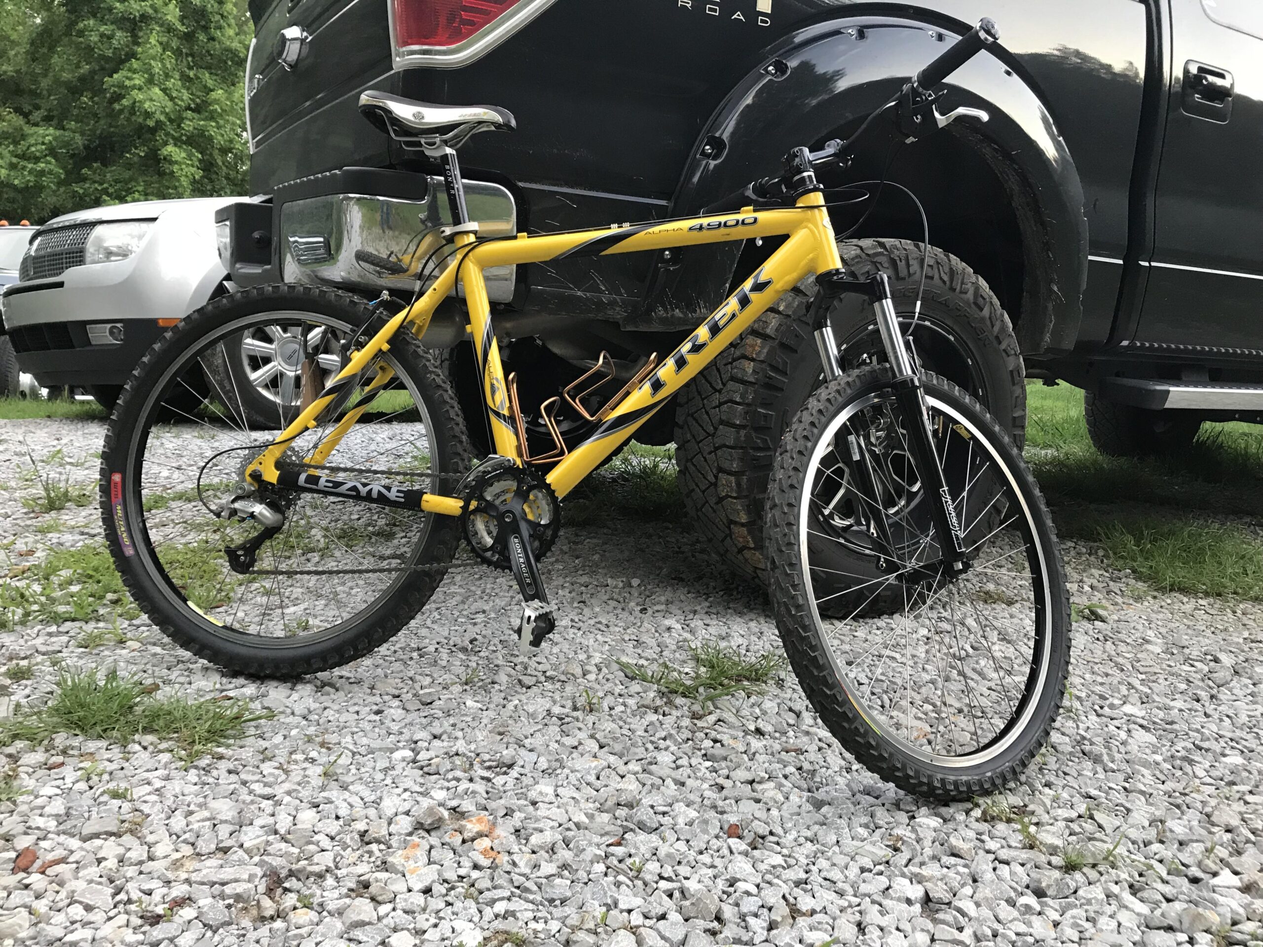 Trek 4900: A yellow Trek mountain bike is parked on a gravel surface next to two vehicles, one of which is a black truck. The bike has a sturdy frame and visible features like thick tires and a prominent gear system. Surrounding the bike, patches of grass and gravel are visible. The background shows a wooded area.