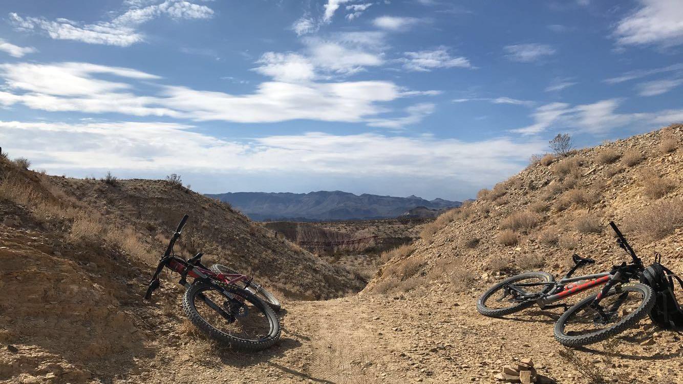 Two mountain bikes are resting on a dirt path surrounded by rocky terrain and sparse vegetation. In the background, rolling hills and mountains are visible under a partly cloudy sky. Big Bend State Park mountain bike trail.