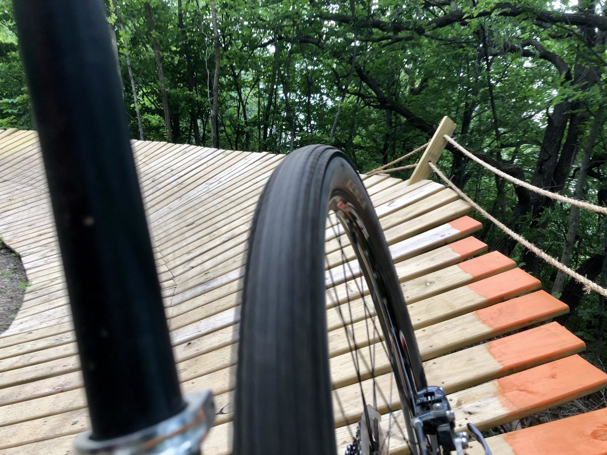 A close-up view of a bicycle tire on a wooden path that curves through a green forested area. The path has alternating orange and natural wood colors and is elevated, with trees visible in the background. Bertram Chain of Lakes Trail mountain bike trail.
