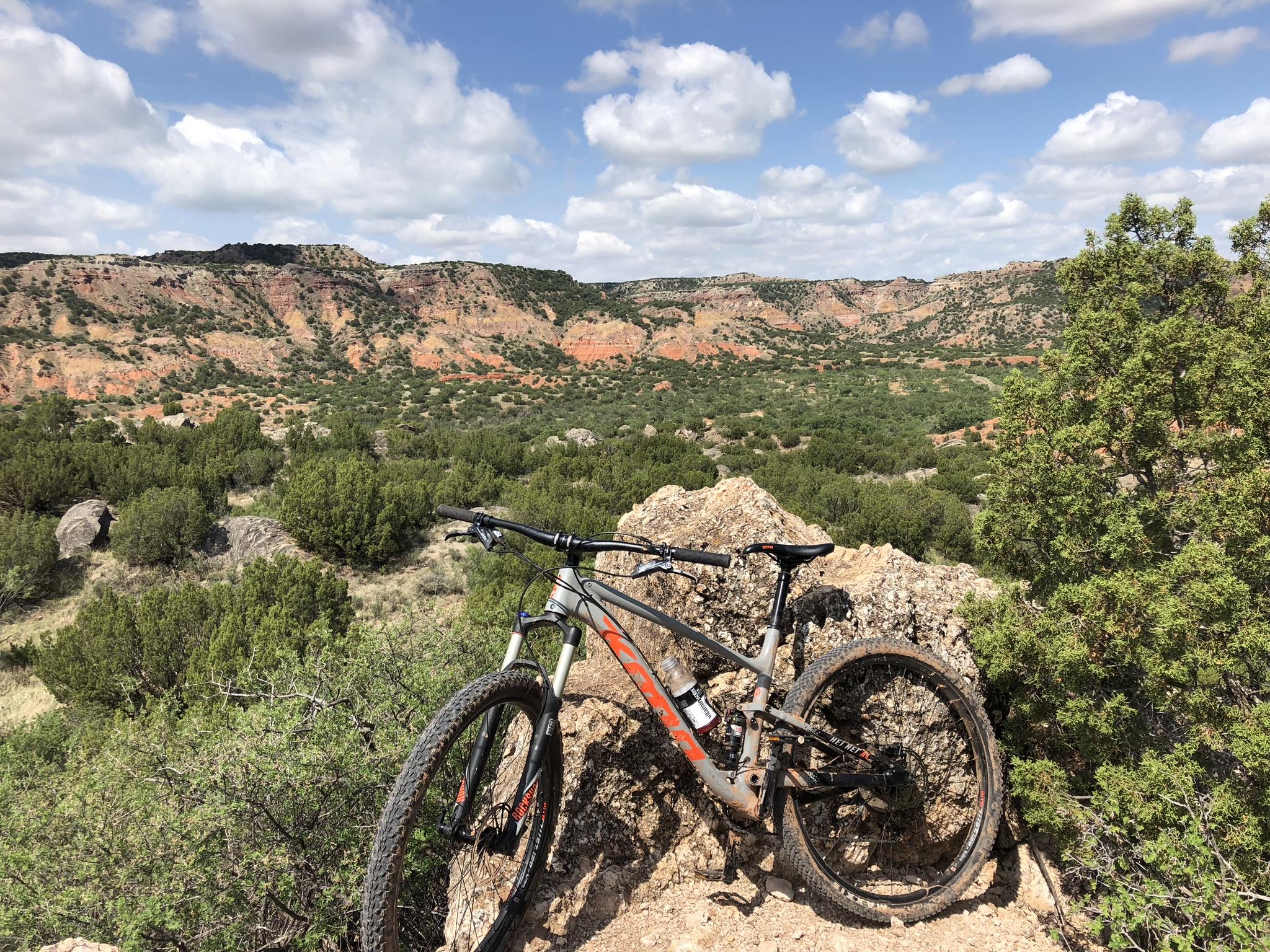 Mountain bike resting on a rocky outcrop with a scenic view of red rock formations and green shrubbery in the background, under a partly cloudy blue sky. Palo Duro Canyon mountain bike trail.