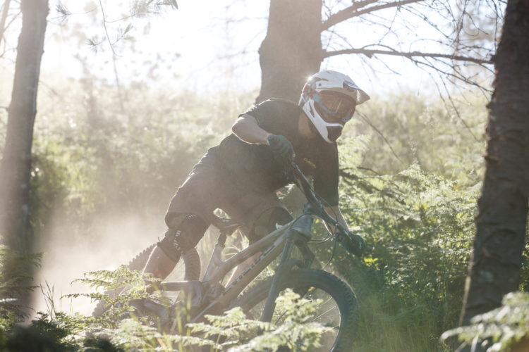 A mountain biker navigates a dusty trail through a forest, showcasing dynamic movement as they corner sharply. The scene is illuminated by soft sunlight filtering through the trees, highlighting the natural vegetation and creating a vibrant atmosphere of adventure. The biker is wearing protective gear including a helmet and gloves.
