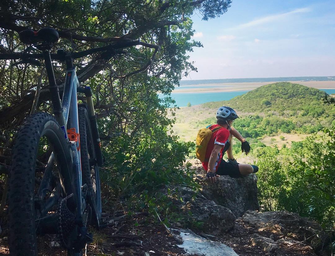 A mountain biker resting on a rock overlooks a scenic view of a lake and rolling hills, framed by greenery. A mountain bike is leaned against a nearby bush, highlighting the outdoor adventure. Dana Peak mountain bike trail.