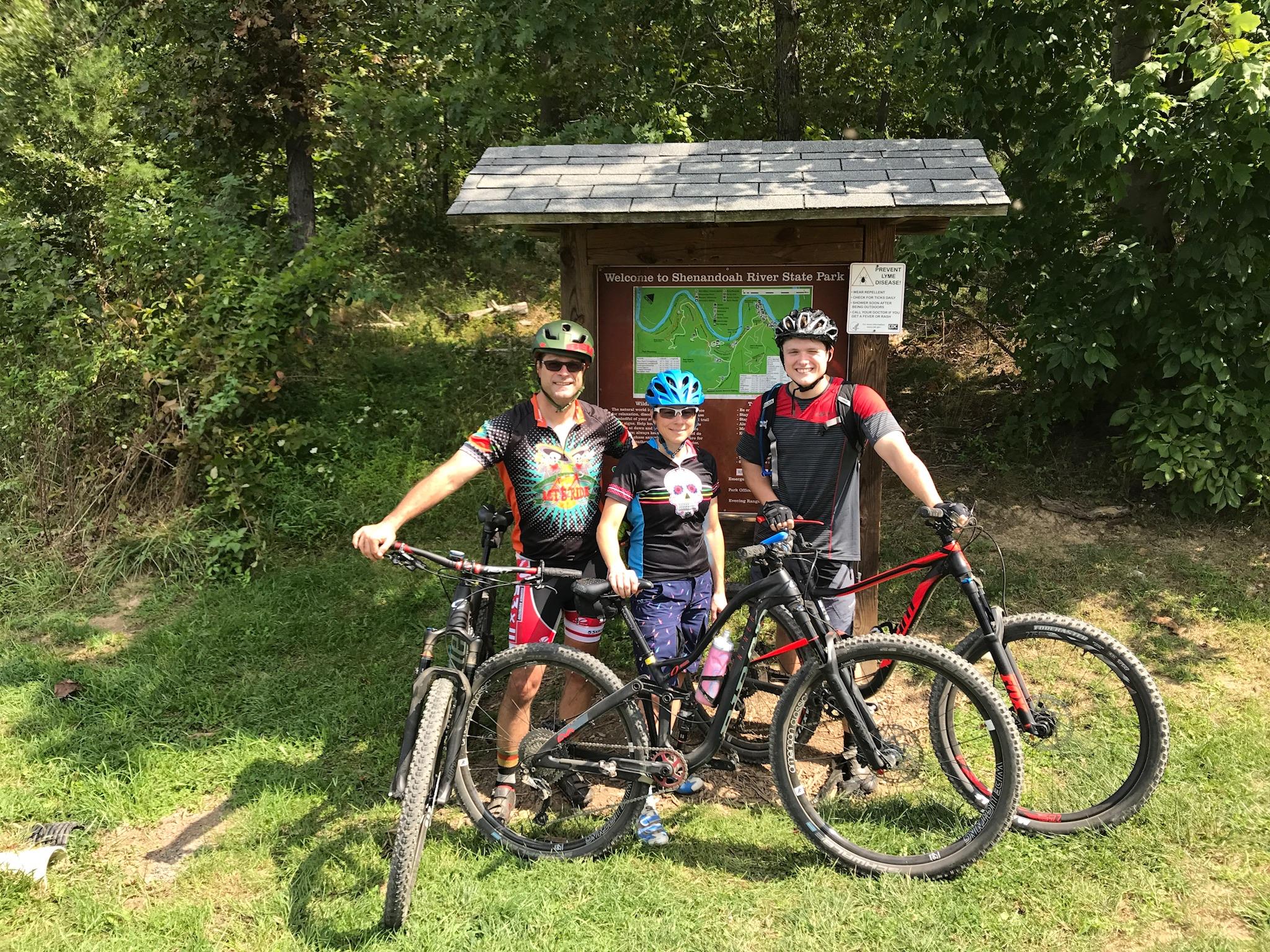 Niner Jet 9 Carbon: Three mountain bikers pose for a photo in front of a park sign at Shenandoah River State Park. They are dressed in colorful cycling attire and wearing helmets. Two adult men and a child stand with their bikes on a grassy area surrounded by trees. The sign behind them features a map of the park.