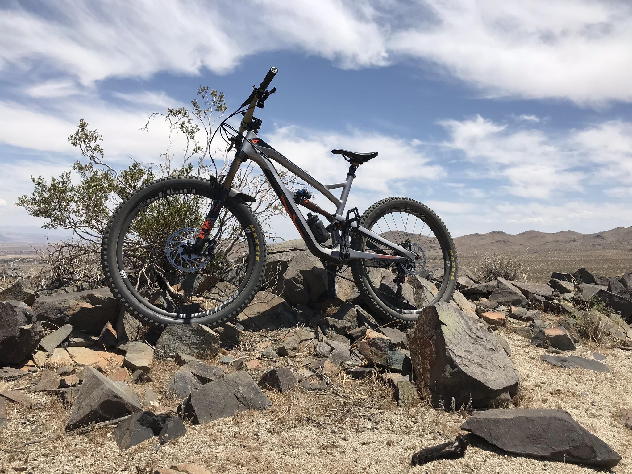YT Industries YT Industries Jeffsy CF Pro Race 27: A mountain bike resting on a pile of rocks with a barren landscape and a cloudy sky in the background. The bike is gray with orange accents, positioned at an angle that highlights its wheels and frame. Sparse vegetation can be seen nearby, and the distant hills blend into the horizon.