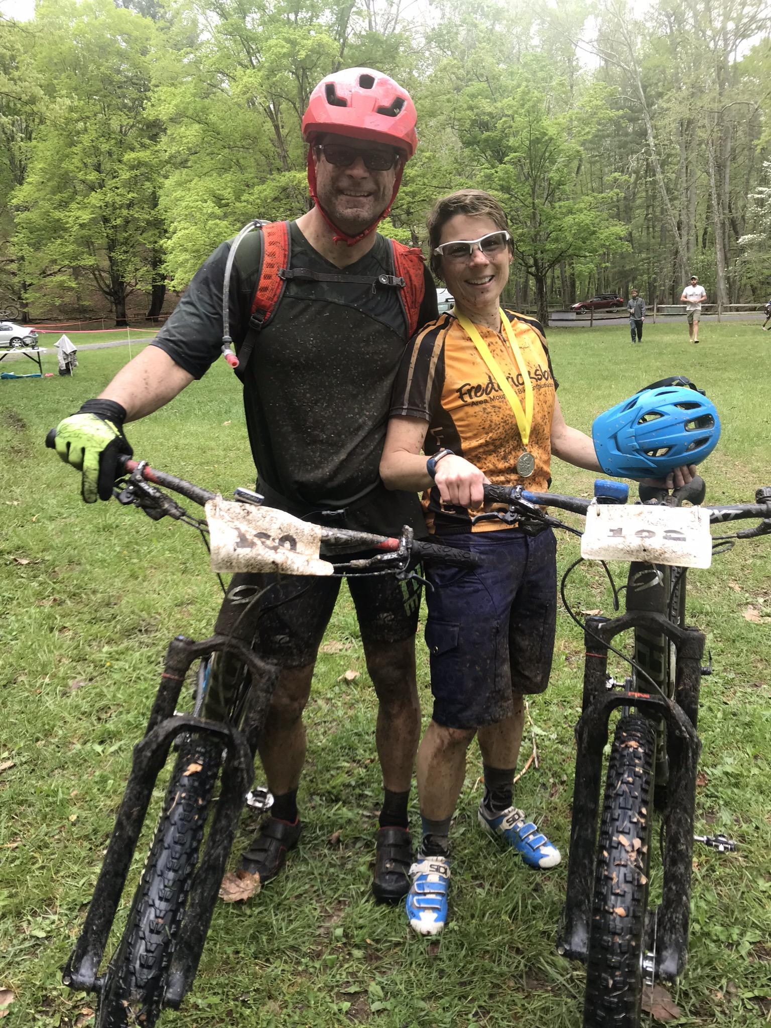 Niner Jet 9 Carbon: A man and a woman stand together in a grassy area, both covered in mud from biking. The man, wearing a red helmet and black clothing with a hydration pack, smiles as he holds his bike. The woman, wearing sunglasses and a brown and yellow cycling jersey with a medal around her neck, also holds her bike and has a blue helmet tucked under her arm. In the background, trees and a few people can be seen.