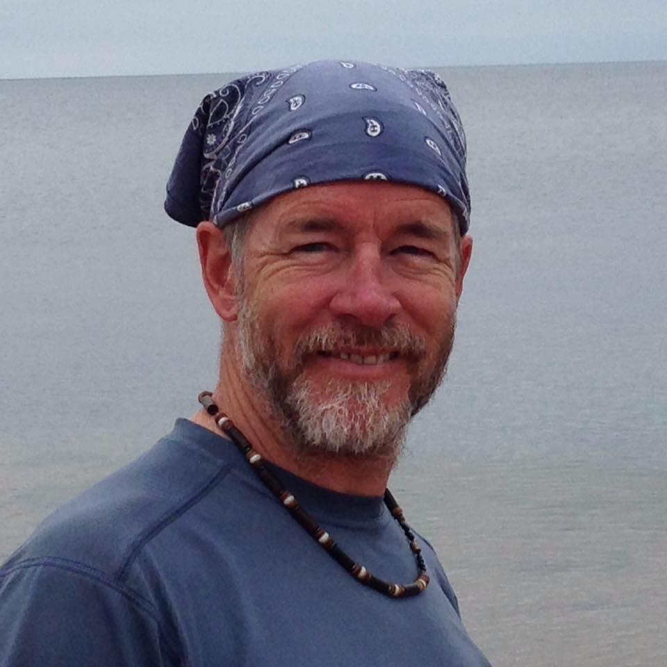 Specialized Stumpjumper FSR: A smiling man with a beard, wearing a blue bandana and a gray shirt, stands by the water with a calm sea in the background. He has a beaded necklace and is looking directly at the camera.