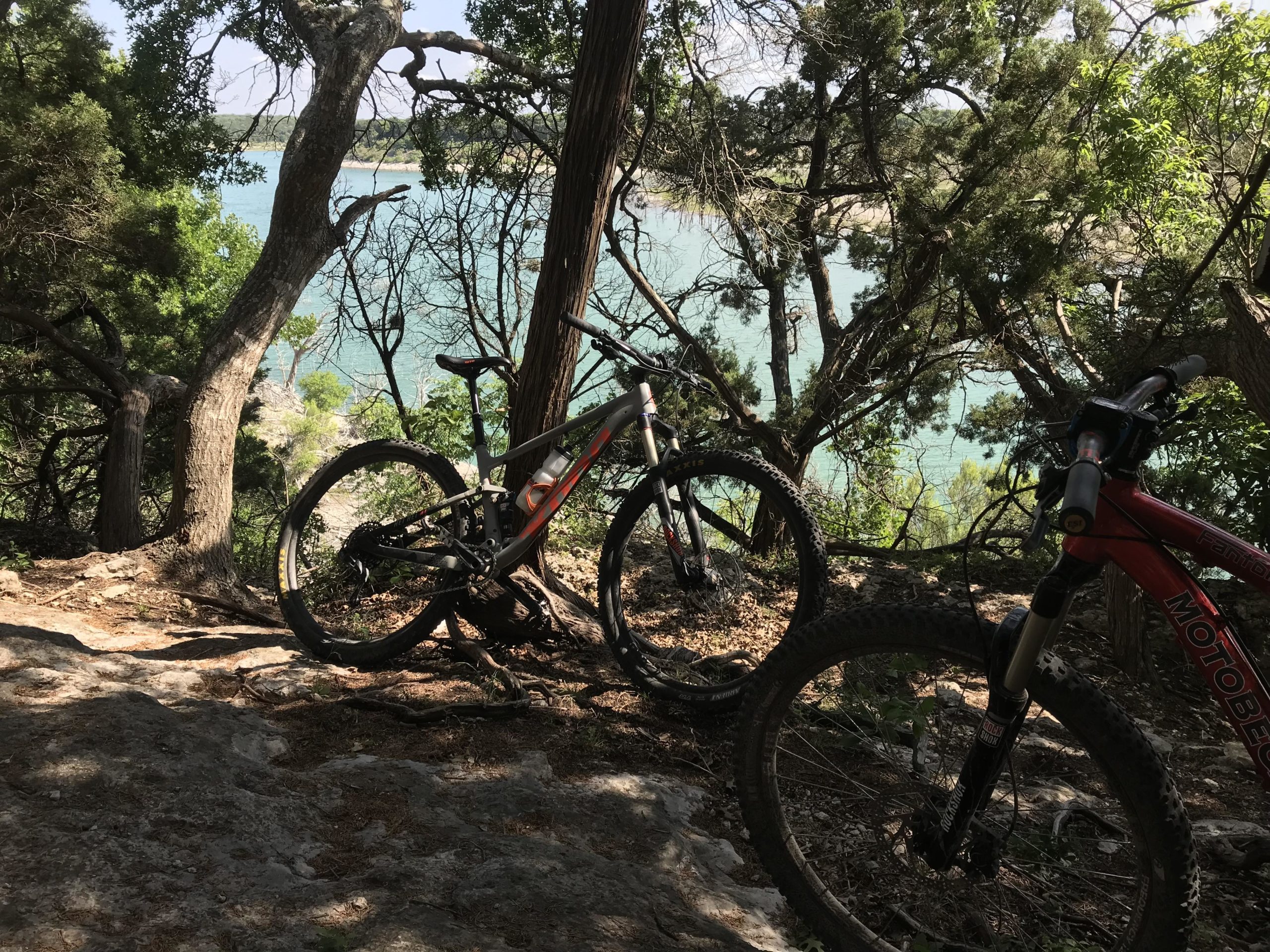 A scenic view of a lake surrounded by trees, with two mountain bikes resting on a rocky trail in the foreground. The water is a vibrant blue, visible through the foliage, creating a peaceful outdoor setting for biking or hiking. Goodwater Trail mountain bike trail.