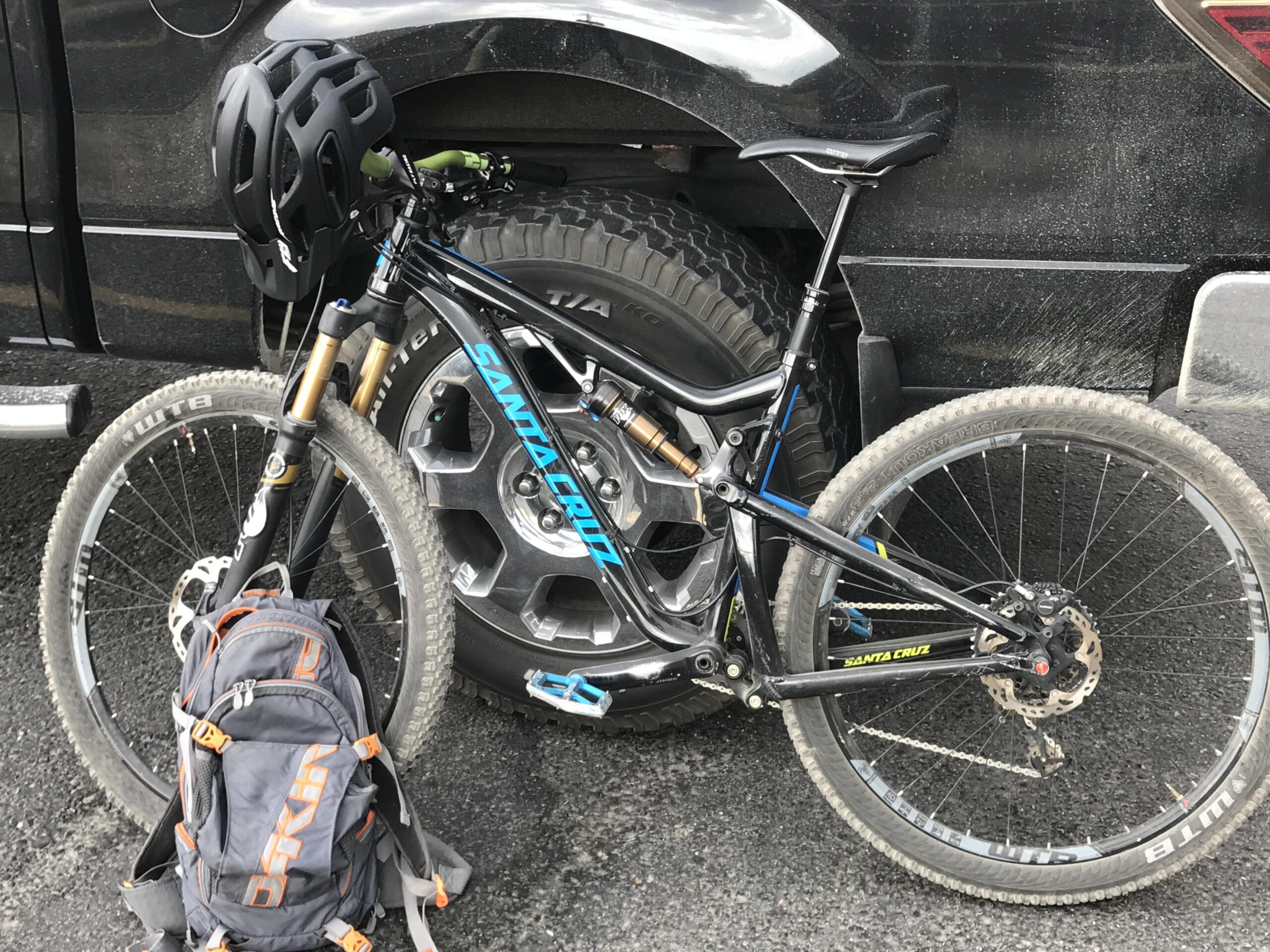 Santa Cruz Bronson: A mountain bike leaning against the wheel of a black truck, with a black helmet resting on the handlebars. A gray backpack with orange accents is positioned on the ground next to the bike. The background shows the truck's side and a textured road surface.