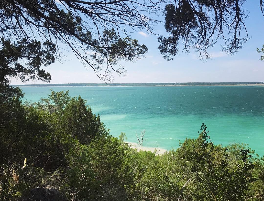 A scenic view of turquoise waters framed by green trees, with a clear blue sky overhead and a distant shoreline in the background. Dana Peak mountain bike trail.