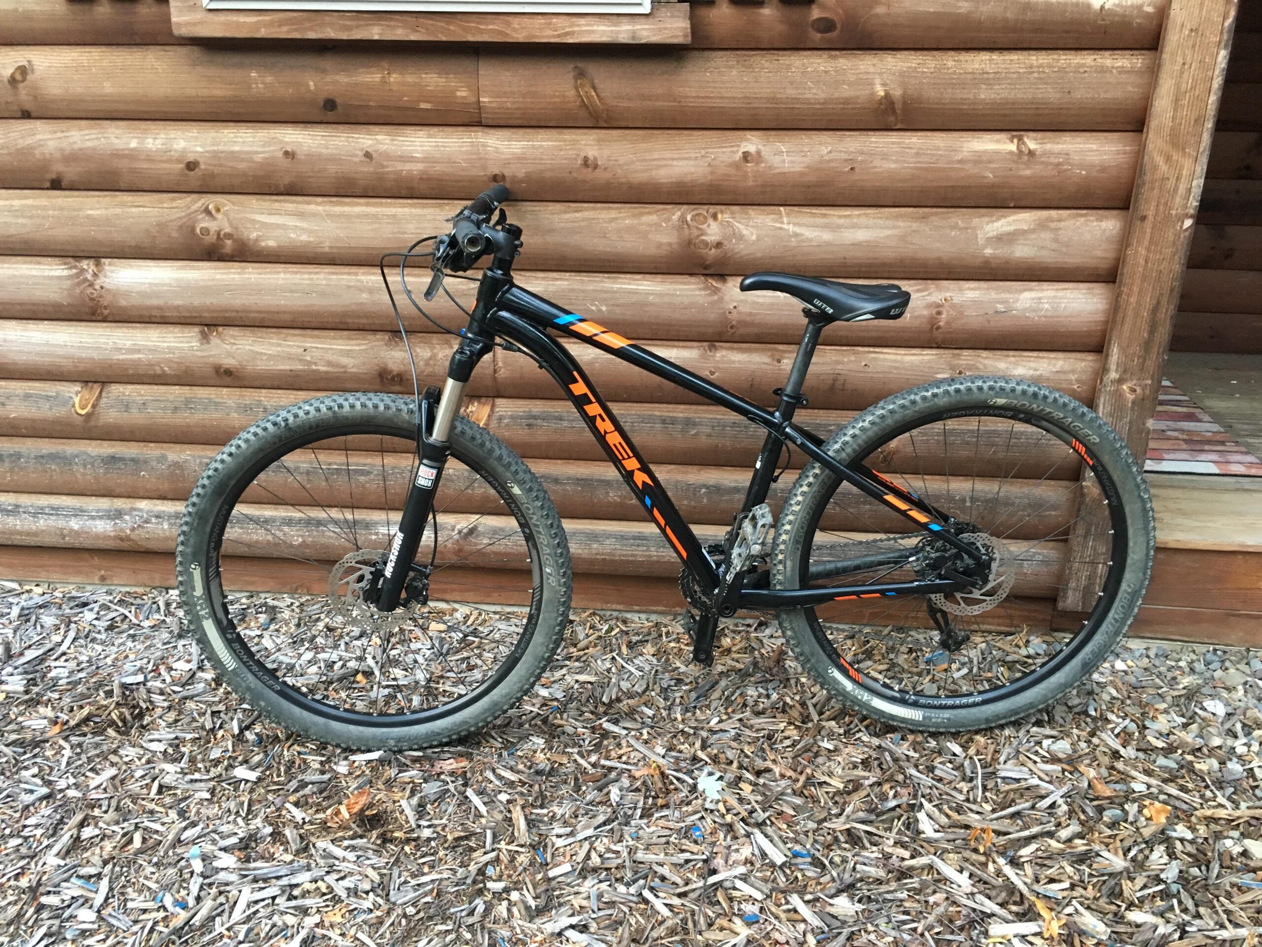 Trek Xcaliber 8: A black and orange mountain bike is leaning against a wooden log cabin. The bike features thick tires, a suspension fork, and a simple handlebar setup. The ground is covered with wood chips, and the background shows the log cabin's wall and a small porch area.