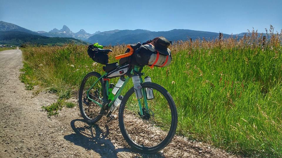 A green bicycle parked on a dirt road beside tall grass, with mountains in the background under a clear blue sky. The bicycle is equipped with bags and water bottles, suggesting it is set up for a cycling adventure or tour.