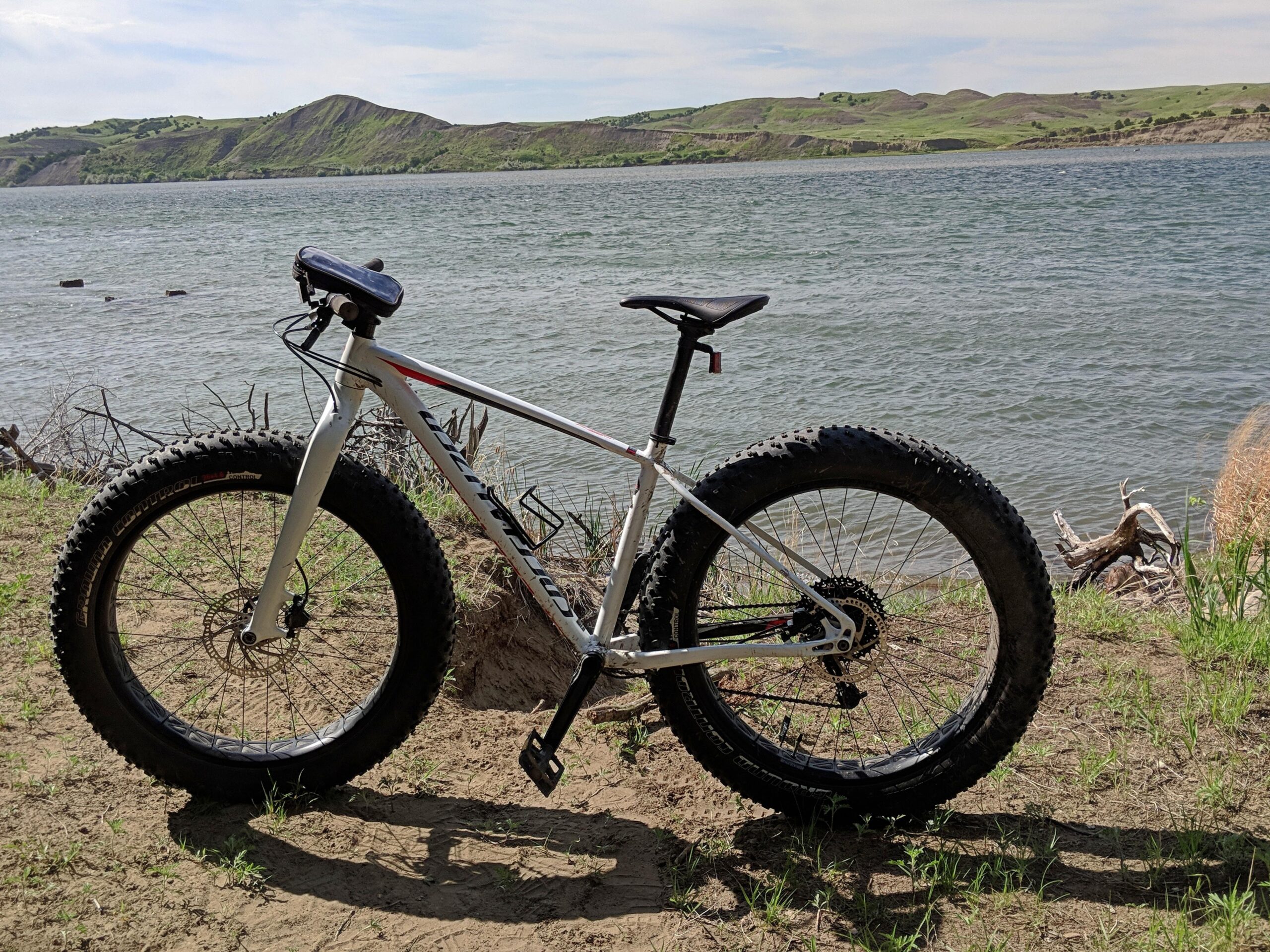 Specialized Fatboy: A fat tire bike parked on a sandy area beside a lake, with green hills in the background under a partly cloudy sky.