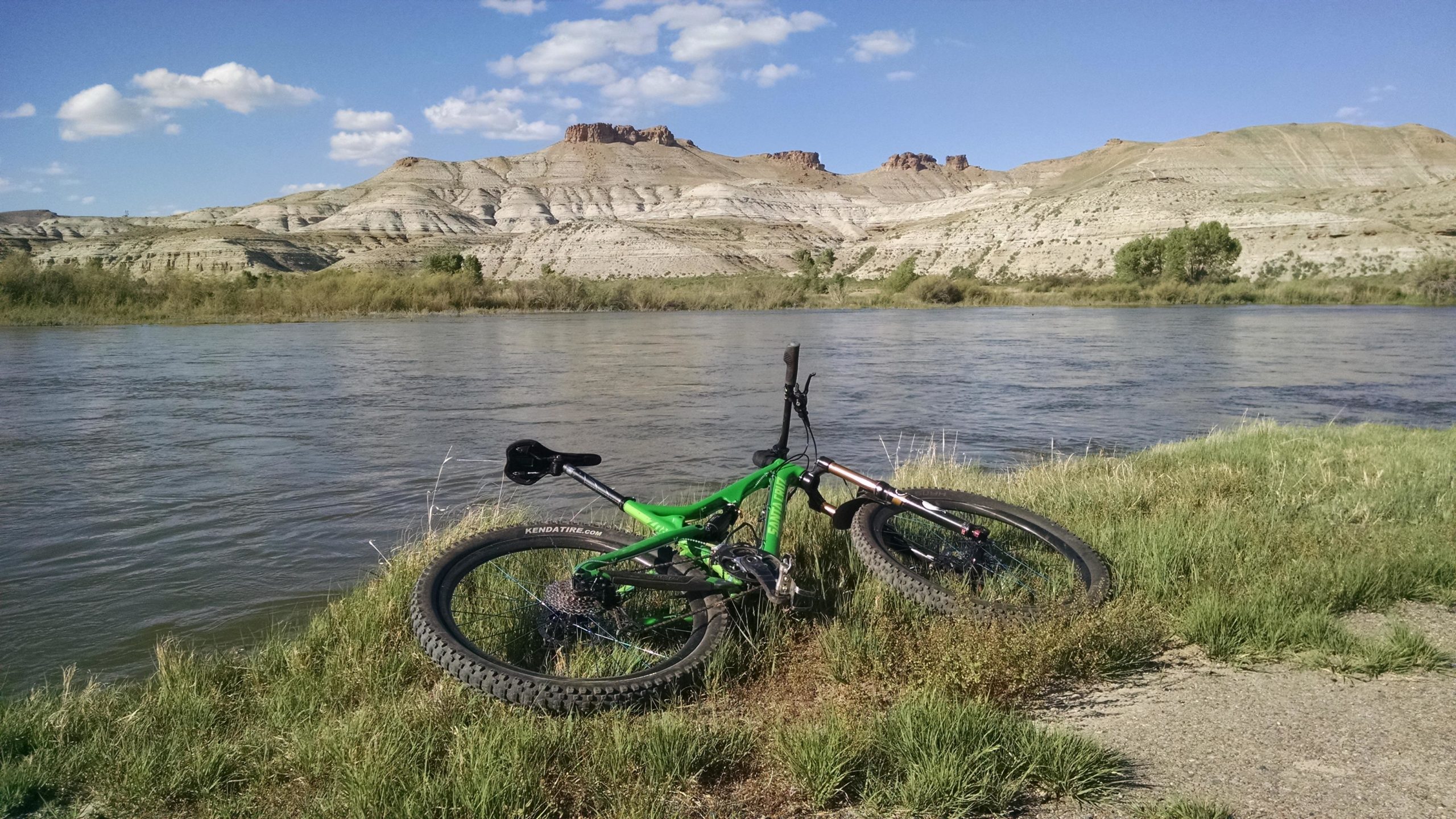 A green mountain bike rests on the grassy shore beside a river, with rocky hills and blue sky in the background. Fluffy clouds are scattered across the sky, and the scene suggests a peaceful outdoor setting ideal for biking and exploring nature. Wilkins Peak Trails mountain bike trail.