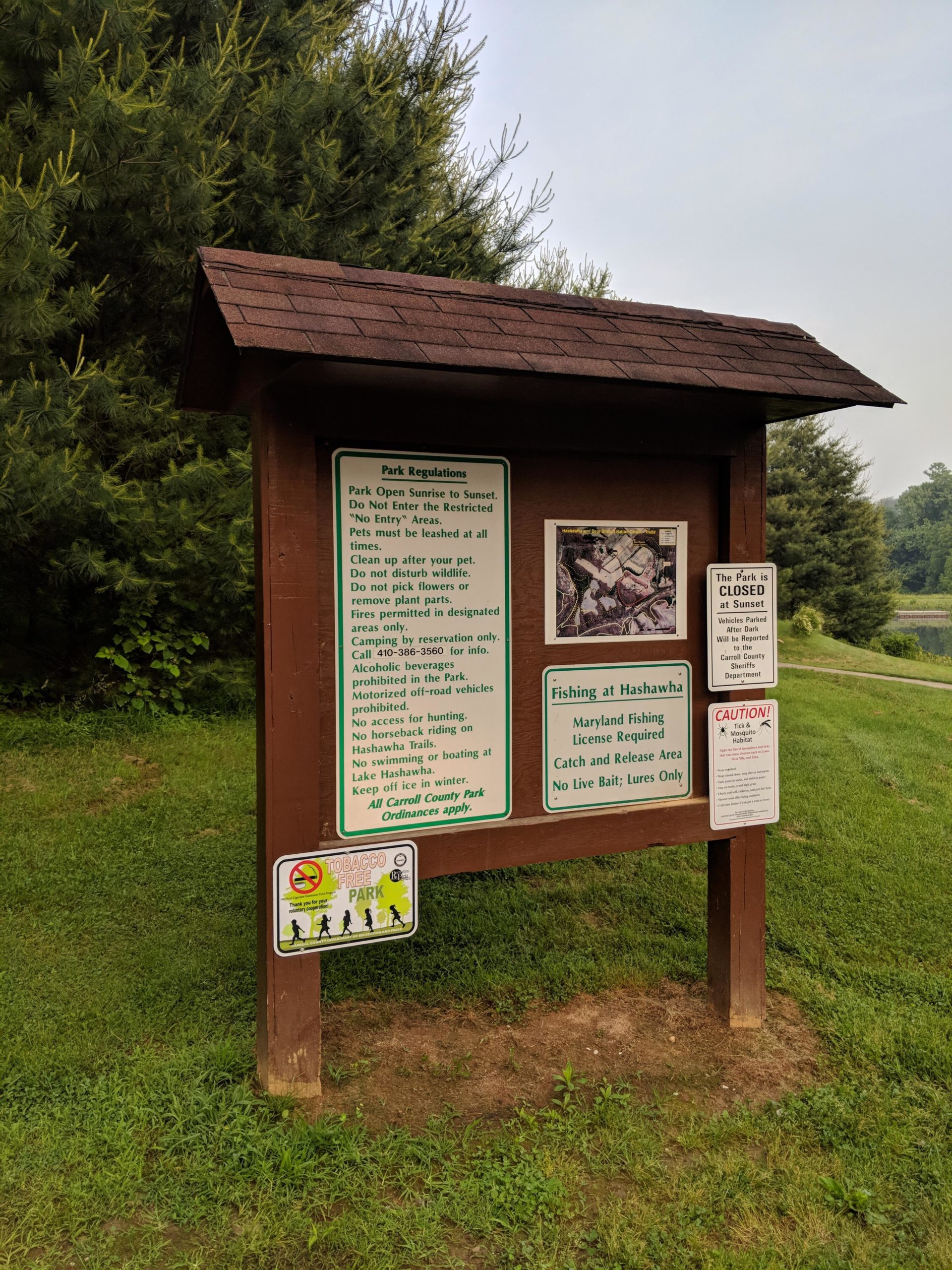 A wooden park regulation sign located in a green outdoor area, surrounded by trees. The sign includes rules for park use, such as operating hours, pet policies, and guidelines for fishing. It also features a map and notices about park restrictions, including no entry areas and a tobacco-free policy. Hashawha mountain bike trail.