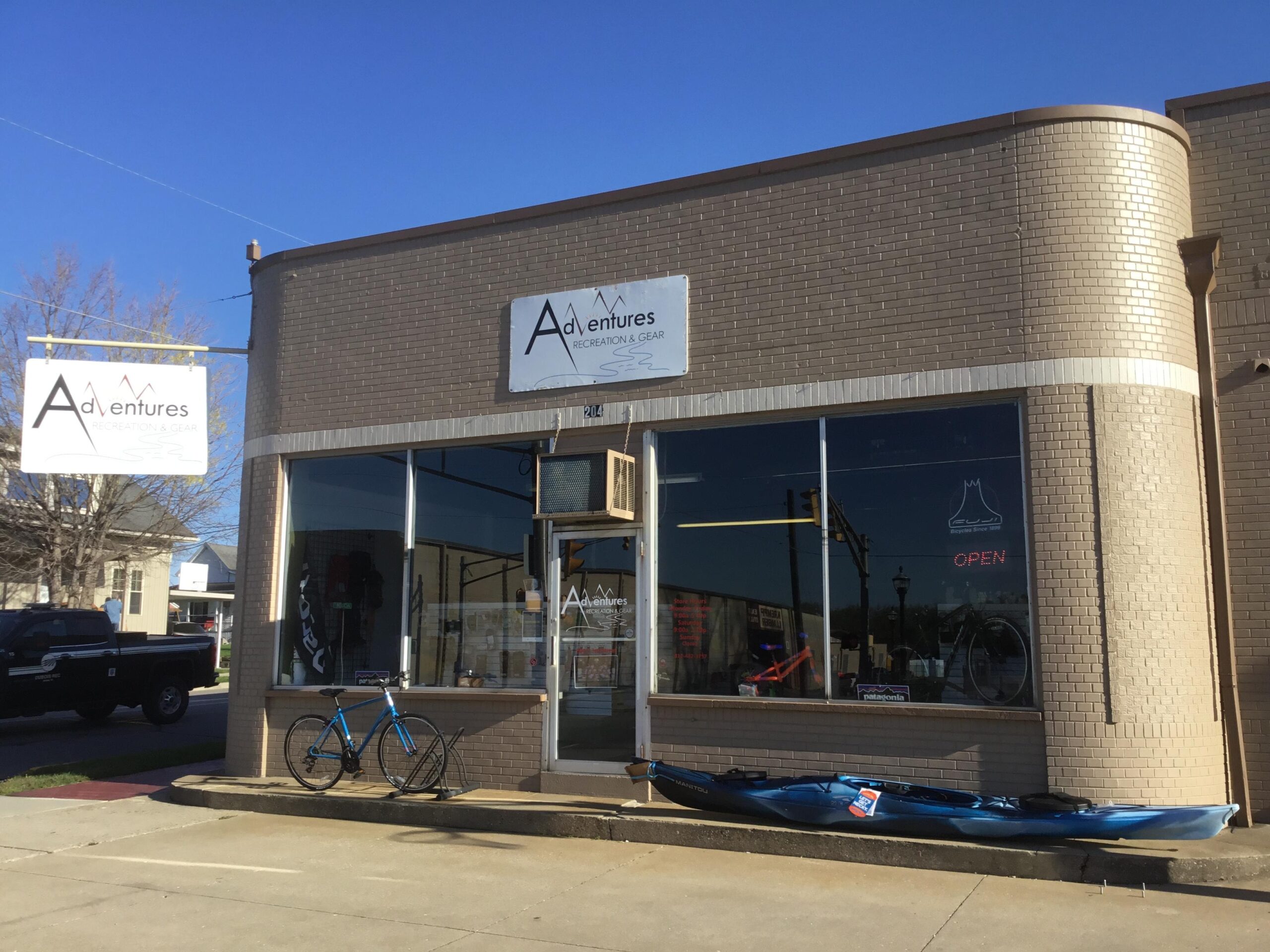 A storefront of a recreation and gear shop named "Adventures," featuring large windows displaying outdoor equipment. A blue bicycle is parked in front, and a kayak is positioned along the sidewalk. The shop has a sign indicating it is open, with clear skies in the background.