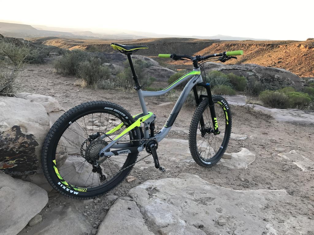 Giant Giant Trance 27.5 2: A modern mountain bike with a gray frame and neon green accents stands on rocky terrain, surrounded by shrubs and distant hills under a sunset sky. The bike features thick tires designed for rough trails and is positioned on large boulders, highlighting its readiness for outdoor adventure.