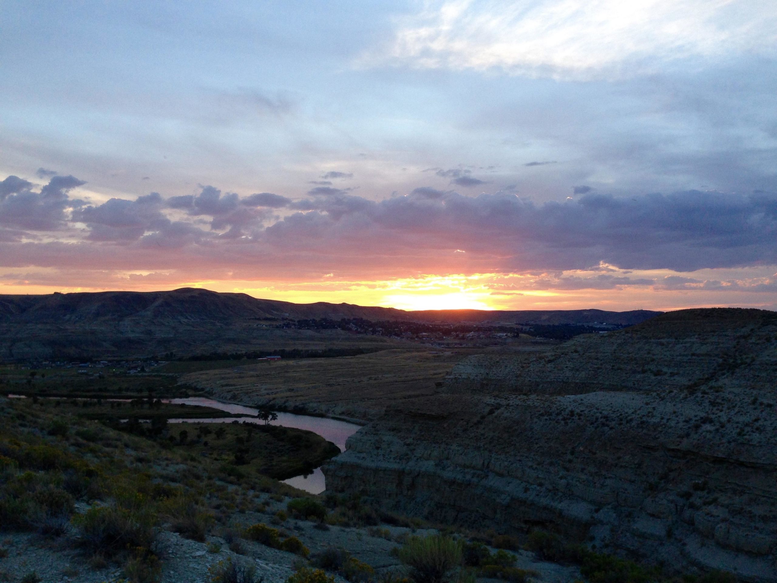Sunset over a rugged landscape featuring rolling hills and a winding river. The sky is filled with clouds painted in shades of orange, pink, and purple as the sun sets on the horizon, casting a warm glow over the terrain. Sparse vegetation is visible in the foreground, with rocky formations creating a textured backdrop. Wilkins Peak Trails mountain bike trail.