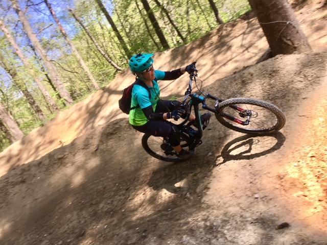 A person riding a mountain bike on a dirt trail in a wooded area, leaning into a turn while wearing a blue helmet and a teal shirt, with a backpack visible. Sunlight filters through the trees, highlighting the natural surroundings. Dual Slalom Course mountain bike trail.
