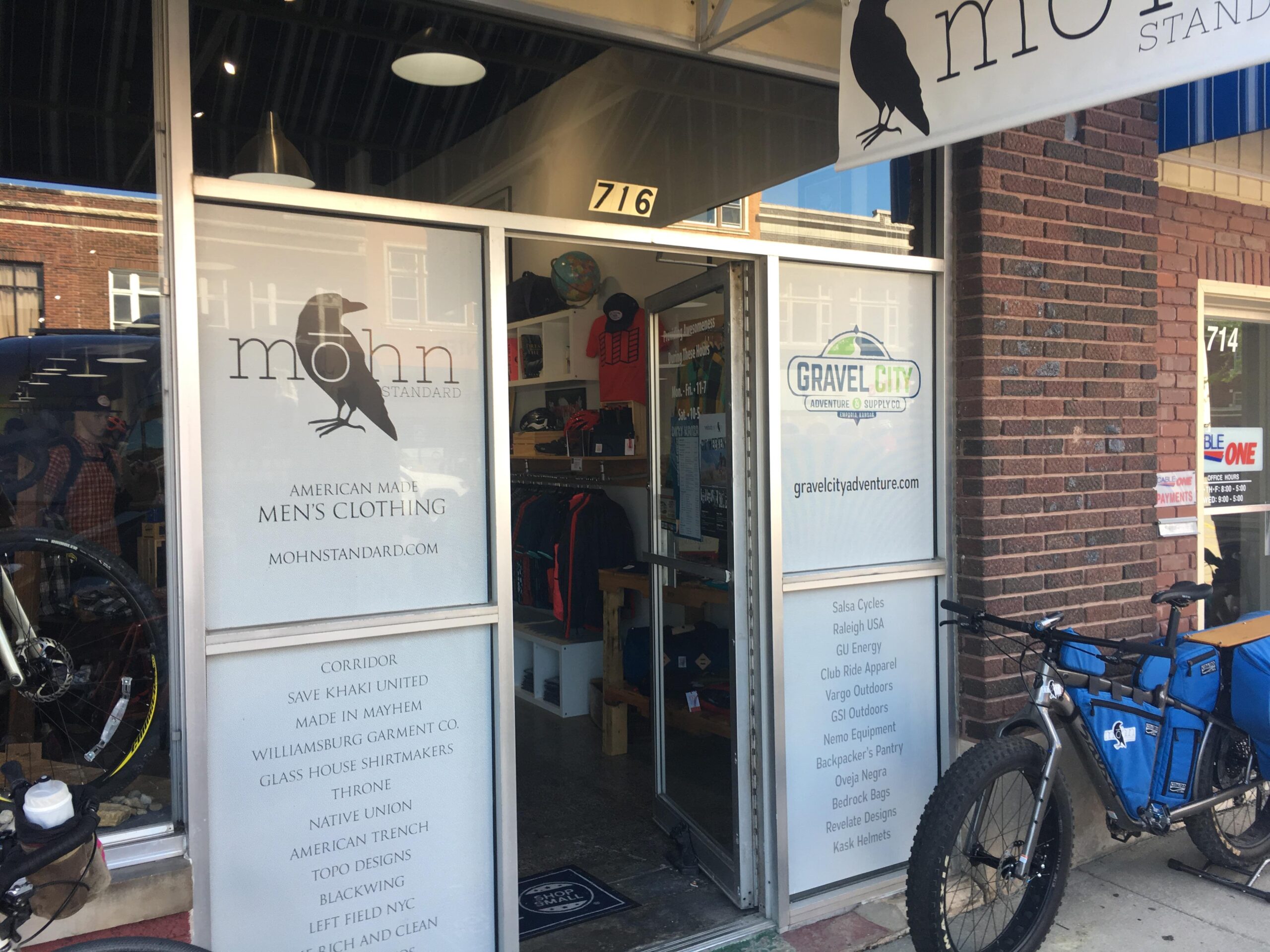 Image of a storefront featuring "mohn" mens clothing, showcasing a modern design with a crow logo. The entrance displays products related to American-made clothing, with a visible bike parked outside. Nearby, a second store named "Gravel City" also shows adventure supplies. The scene captures a vibrant shopping area with brick walls and a clear day.