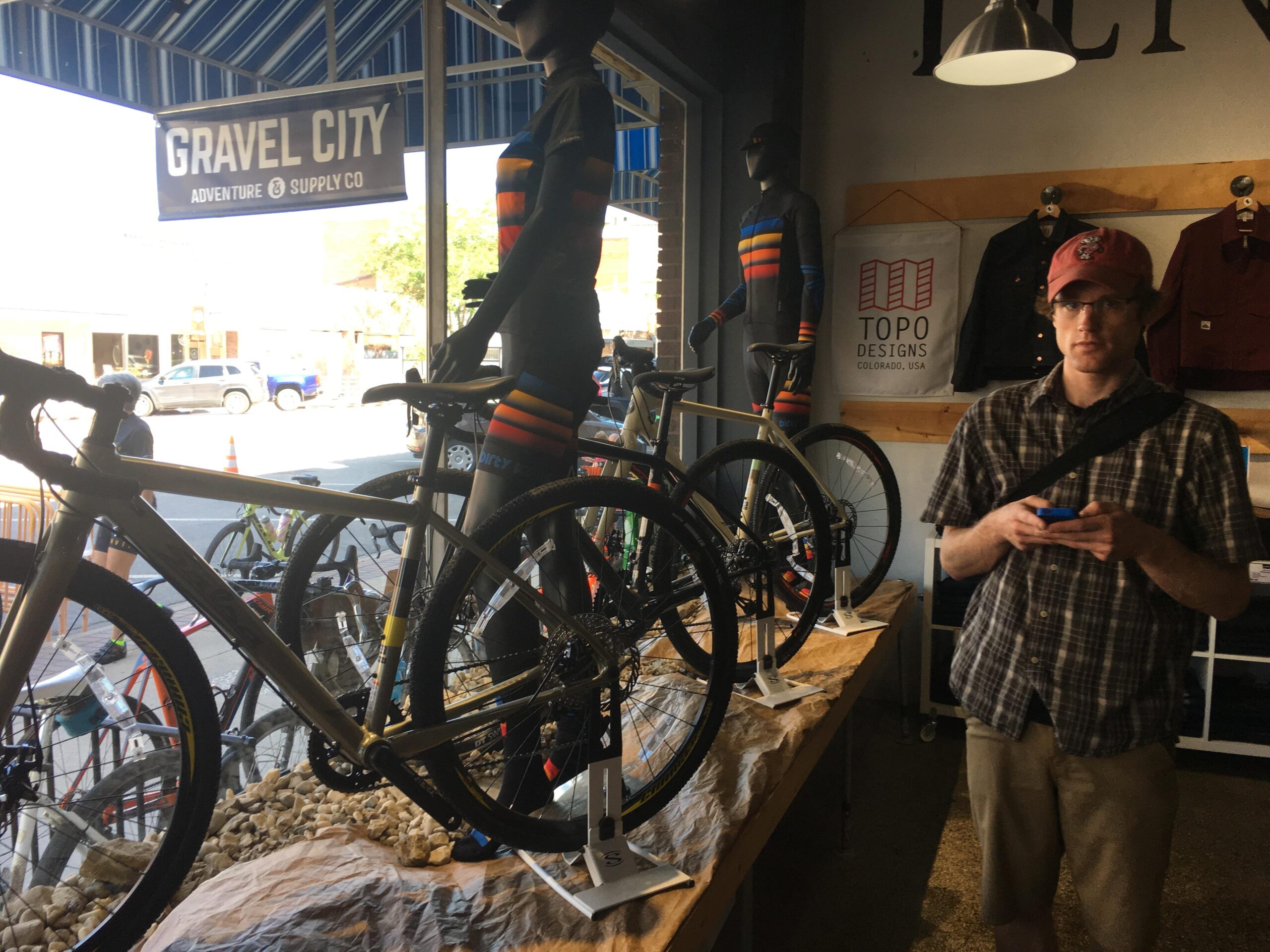A man in a plaid shirt and cap stands in a bike shop, holding a phone and looking at the camera. In the foreground, there are several bicycles displayed on a table, alongside two mannequins wearing colorful cycling kits. The shop features a sign for "Gravel City Adventure Supply Co." in the background, with street scenes visible through large windows.