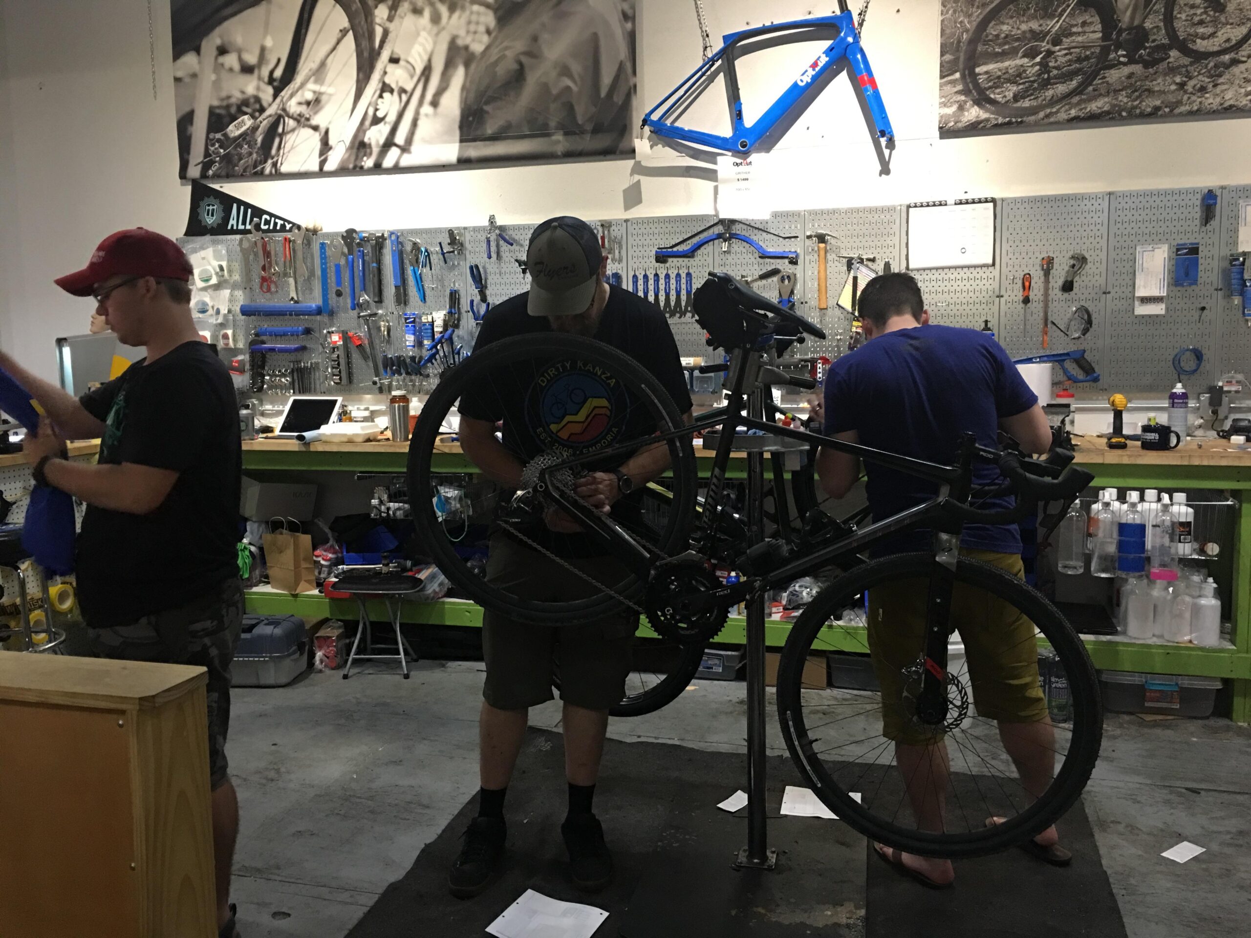 Alt text: Three individuals working in a bicycle repair shop. One person is holding a blue cloth, while another is adjusting a bike on a repair stand. Various tools and parts are visible on the walls behind them, and a blue bike frame hangs above.