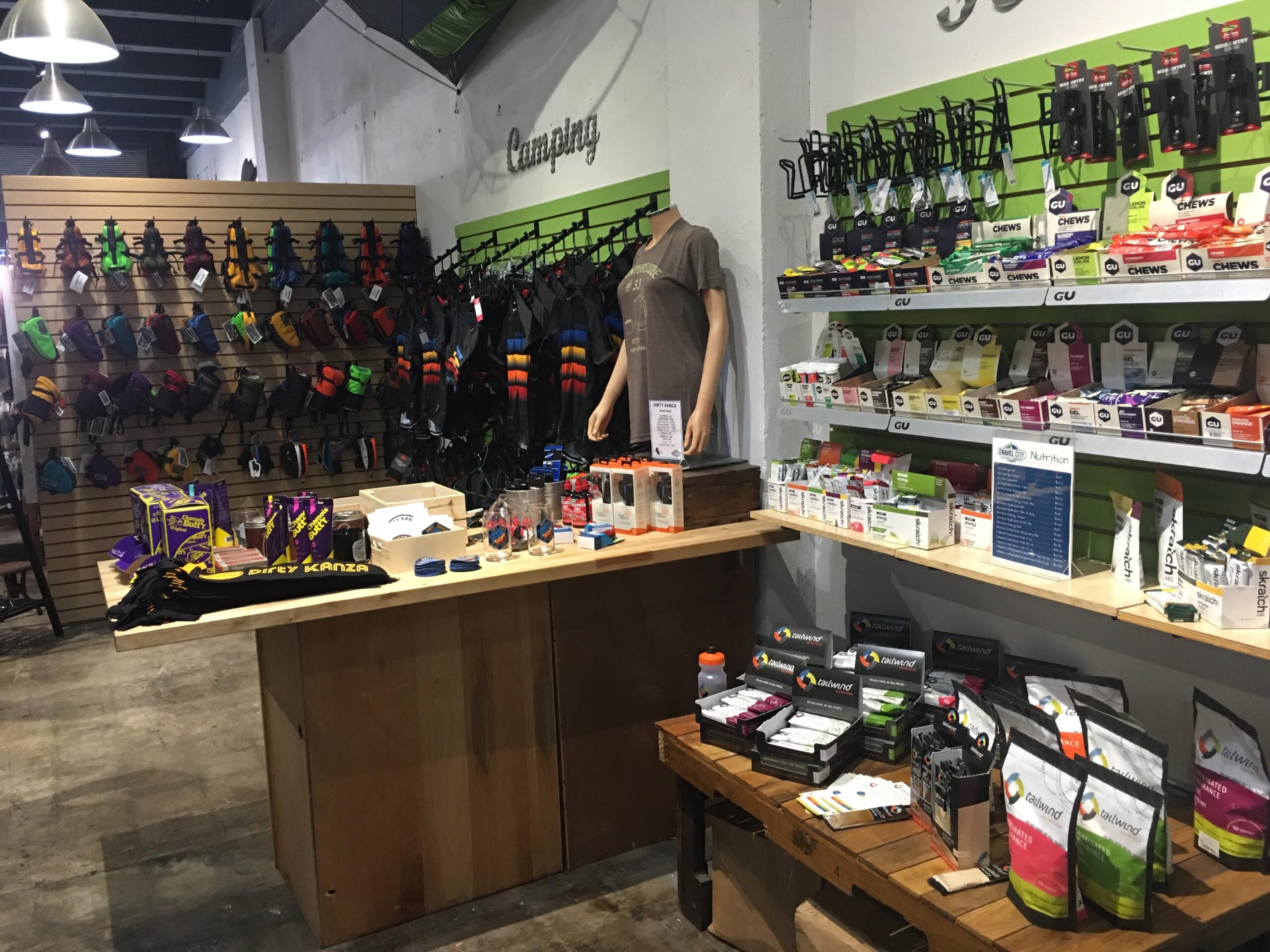 A retail space showcasing a variety of camping and outdoor gear. The left wall features colorful climbing and camping equipment arranged on hooks. A mannequin dressed in outdoor clothing stands prominently, while a wooden table displays various products, including energy snacks, drinks, and branded merchandise. Brightly colored packaging and organized shelves contribute to the vibrant shopping atmosphere.