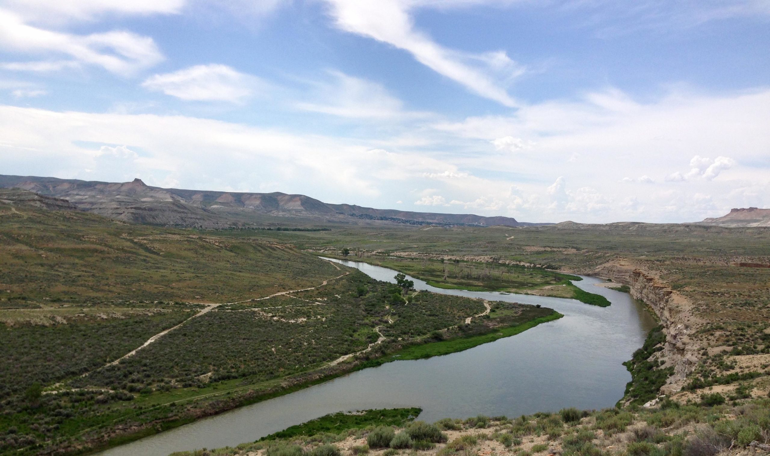 A panoramic view of a winding river surrounded by lush greenery and arid landscapes under a partly cloudy sky. The scene features rolling hills in the background, with a mixture of vegetation and rocky terrain along the riverbanks. Wilkins Peak Trails mountain bike trail.