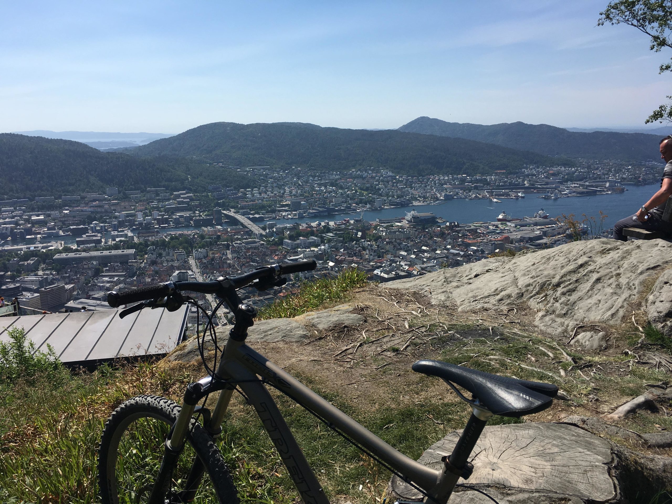 A mountain bike resting on grass near the edge of a rocky overlook, with a panoramic view of a coastal city below, featuring buildings, a bridge, and a harbor. In the background, rolling green hills and clear blue skies are visible. A person is sitting on a rock in the distance, enjoying the scenery. Mount Floyen mountain bike trail.