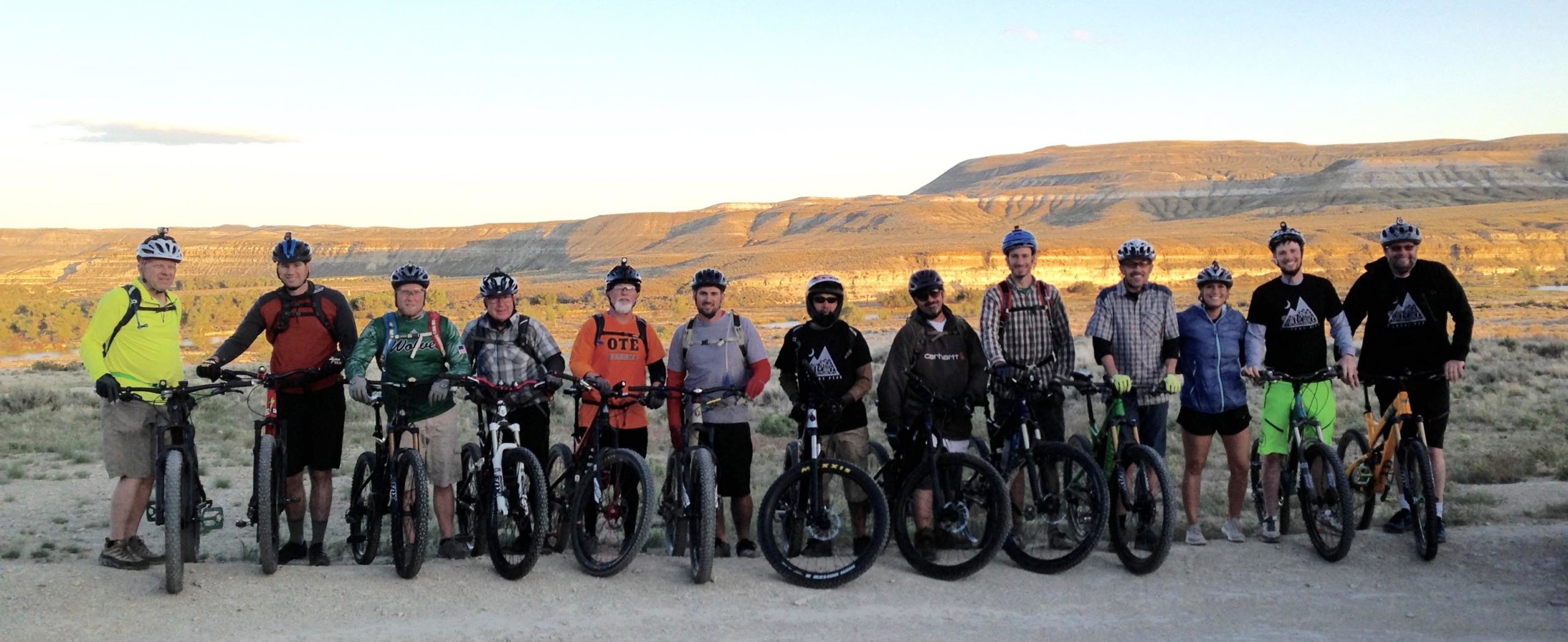 A group of 15 mountain bikers posing for a photo on a dirt path with a scenic landscape in the background, featuring rolling hills and a clear sky. The bikers are wearing helmets and various cycling gear, some with backpacks, and are standing next to their mountain bikes. Wilkins Peak Trails mountain bike trail.