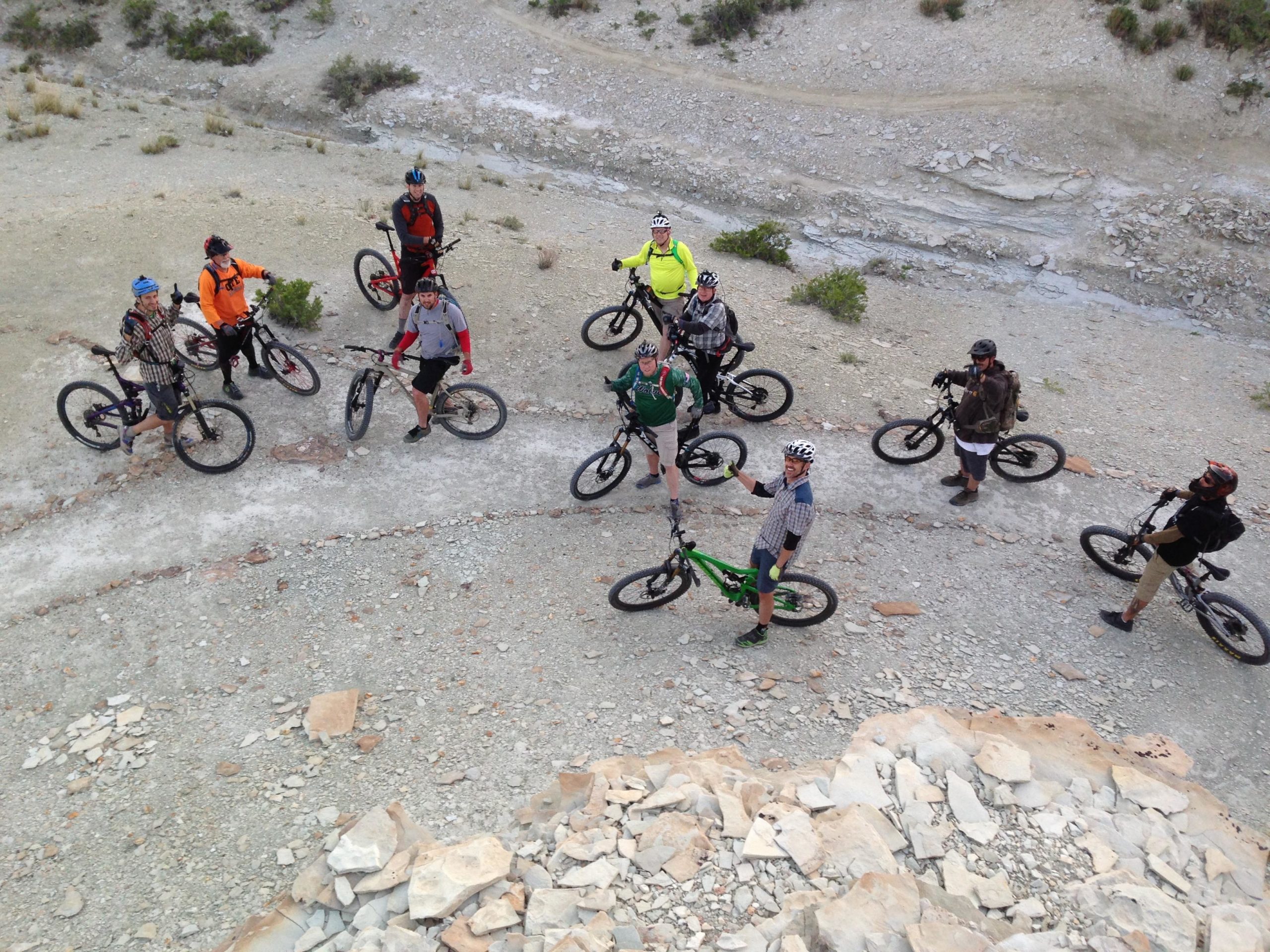 A group of mountain bikers gathered on a rocky terrain, each wearing helmets and biking gear. The riders are standing beside their bikes, with some giving thumbs up and smiling. The landscape features gravel, sparse vegetation, and a winding path. The image captures a sense of camaraderie and outdoor adventure. Wilkins Peak Trails mountain bike trail.