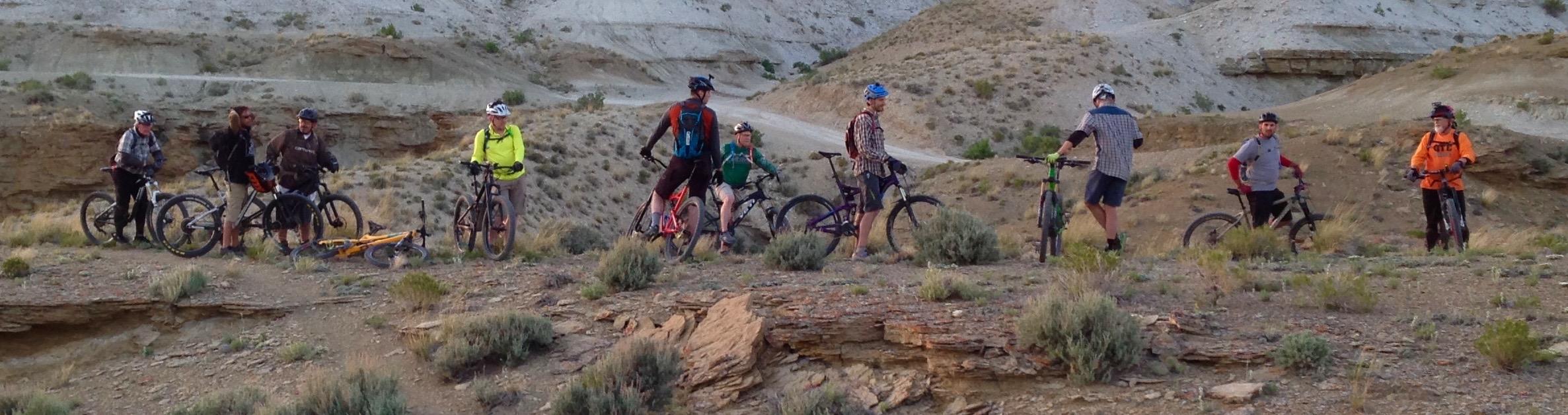 A group of nine mountain bikers stands on a rocky terrain, surrounded by natural landscapes of hills and sparse vegetation. They are dressed in cycling gear and helmets, with various colors represented, while several bikes are positioned nearby. The setting appears to be an outdoor biking trail, showcasing a mix of sunlit and shaded areas. Wilkins Peak Trails mountain bike trail.