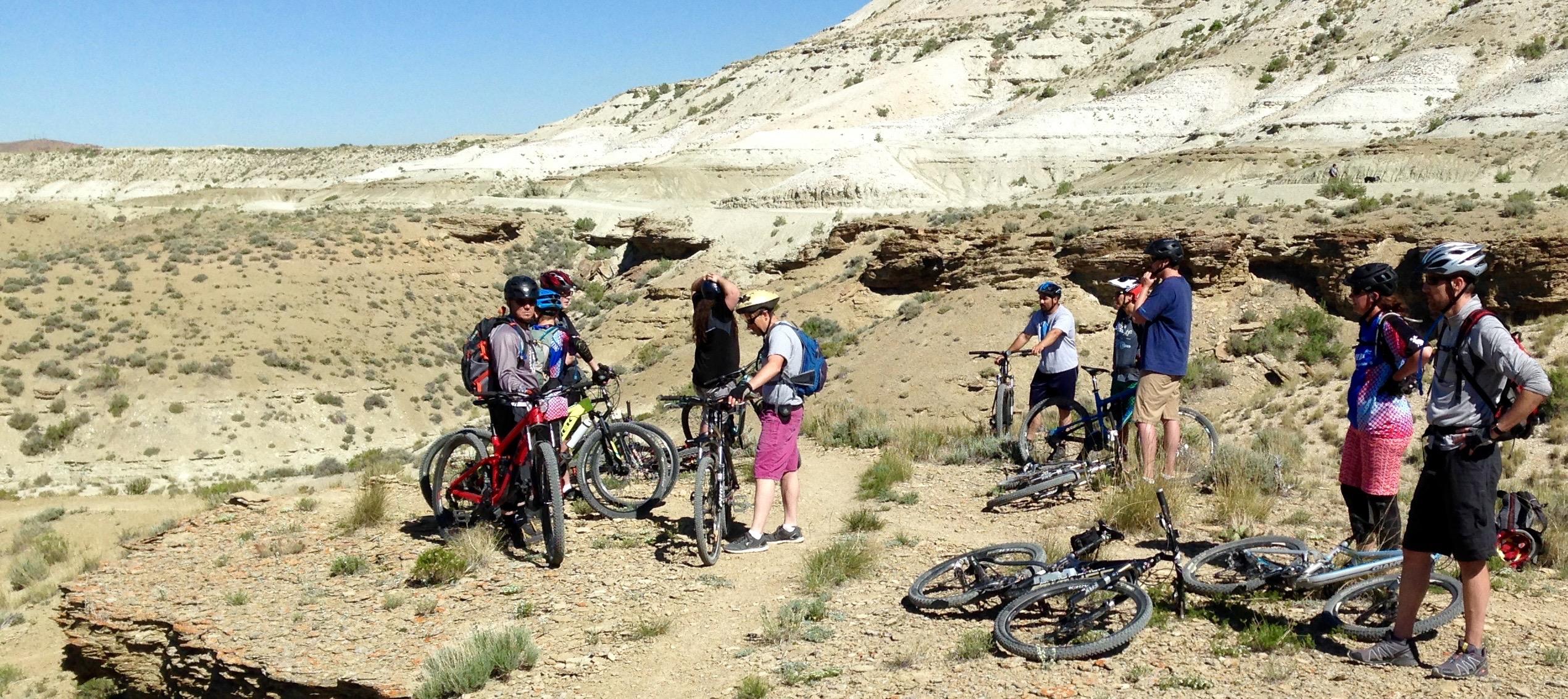 A group of mountain bikers gathered on a rocky ridge in a mountainous desert landscape. Some are checking their bikes, while others are resting or engaging in conversation. The environment features dry, sandy terrain with sparse vegetation and a backdrop of layered rock formations under a clear blue sky. Several bikes are parked nearby, with the riders wearing helmets and casual outdoor clothing. Wilkins Peak Trails mountain bike trail.