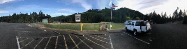 A panoramic view of a parking lot in a grassy area surrounded by forested hills. An American flag flies in the foreground, and there is a sign detailing park information. A white vehicle is parked in the lot, with a few empty spaces visible. The sky is partly cloudy, suggesting a pleasant day. Cooper Creek Trail mountain bike trail.