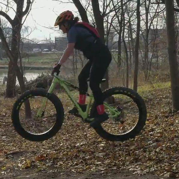Rocky Mountain Blizzard: A person riding a green fat bike performing a jump on a dirt trail surrounded by trees and autumn leaves. The rider is wearing a helmet, gloves, and a backpack, with a body posture indicating an active biking stance. In the background, a river and some urban structures are visible.