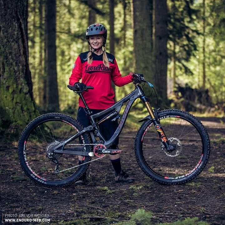 Pivot Mach 6 Carbon: A smiling cyclist wearing a red shirt and a helmet stands beside a mountain bike in a forested area. The bike features thick tires and a sturdy frame, surrounded by tall trees and lush greenery.
