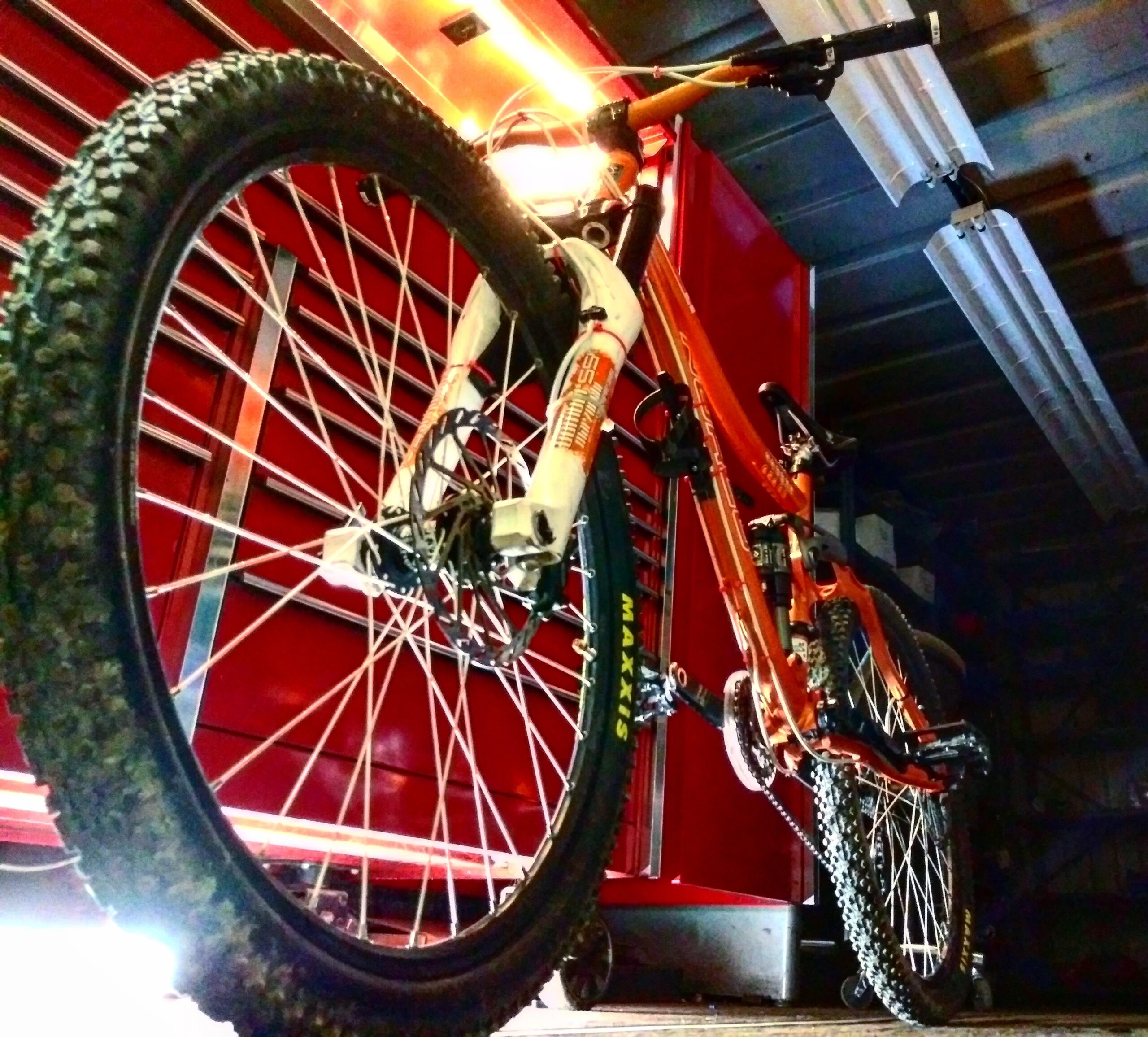 Norco Fluid FS: An off-road mountain bike with thick, knobby tires positioned in a dimly lit garage. The bike features a vibrant orange frame and white suspension fork, with tools and a tool cabinet visible in the background.