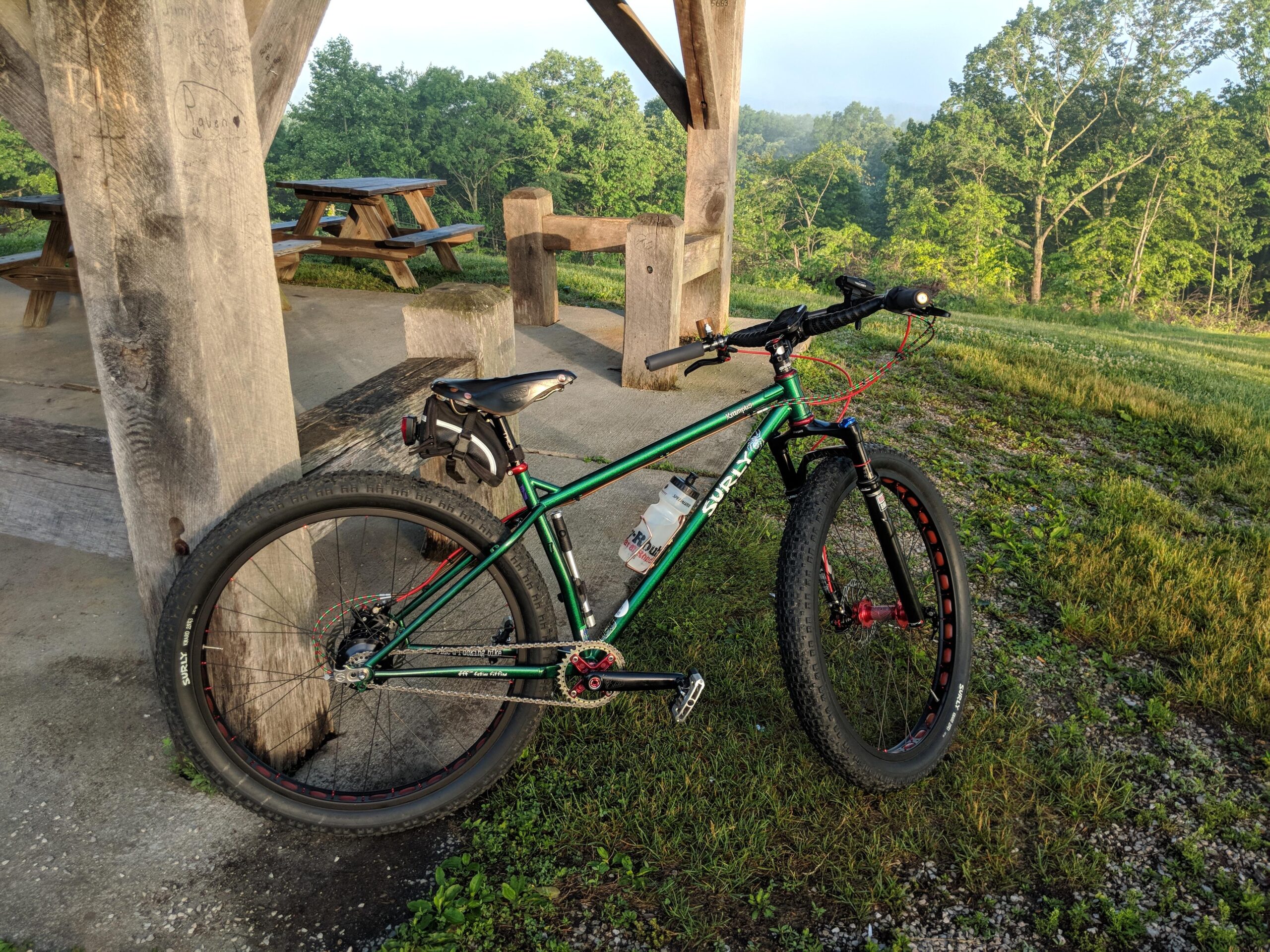 Surly Krampus: A green and black mountain bike with large tires parked near a wooden pavilion. The background features trees and grassy areas, suggesting an outdoor setting. The bike has a water bottle attached and is positioned at the edge of a gravel path.