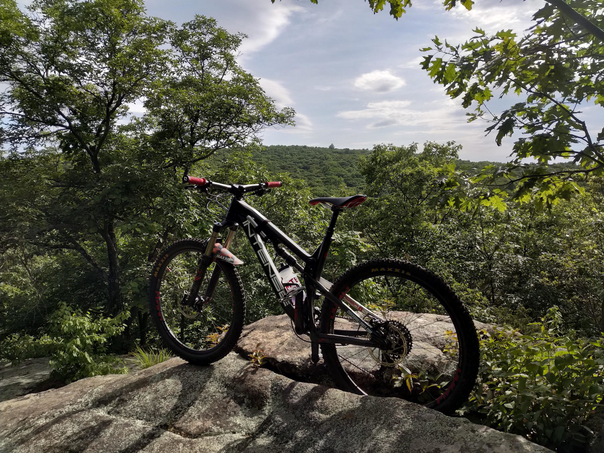 A mountain bike resting on a large rock, surrounded by lush green trees and a scenic view of rolling hills under a partly cloudy sky. Allamuchy State Park-North mountain bike trail.