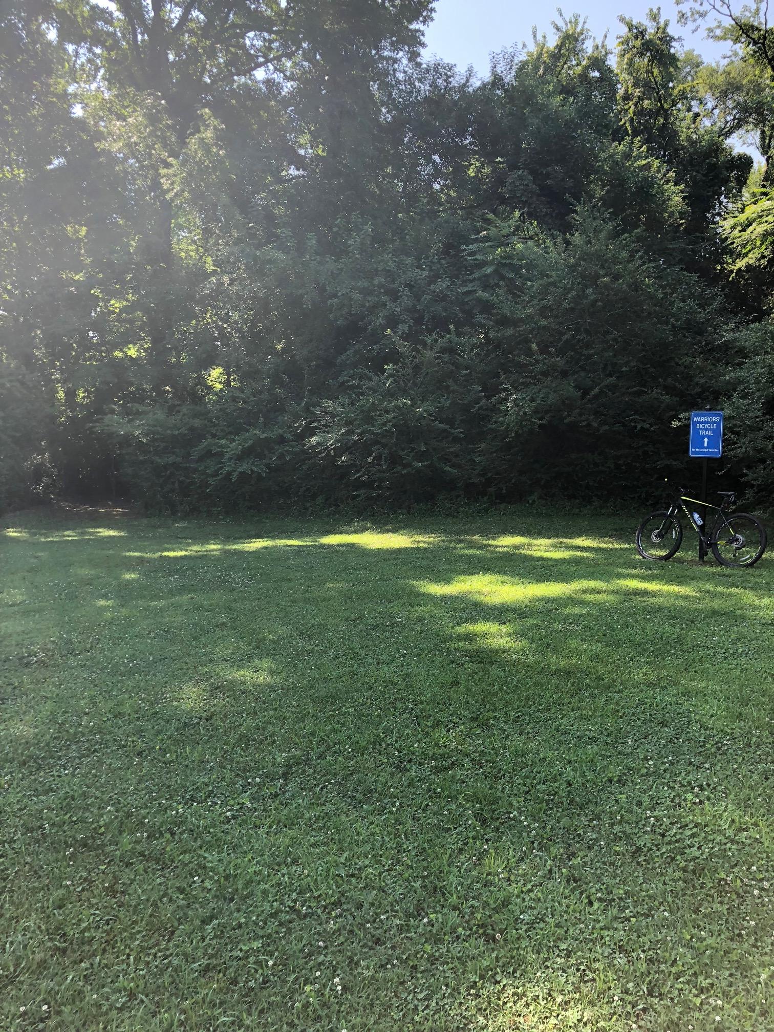 A sunny scene of a grassy open area bordered by trees, featuring a bicycle parked next to a sign that reads "Waltersford Trail," indicating the start of a hiking or biking trail. Wessington Warrior Trail mountain bike trail.