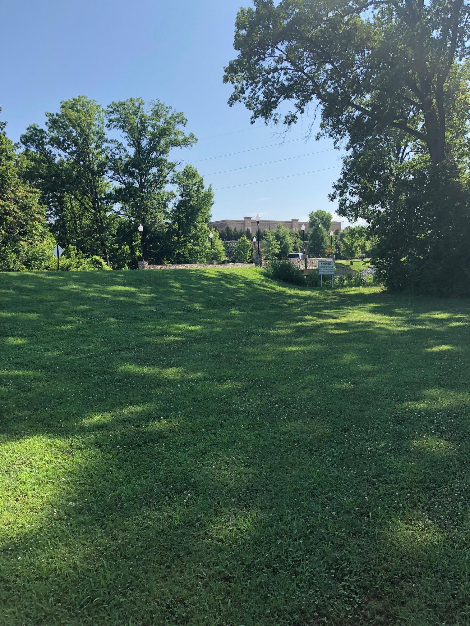 A sunny day view of a grassy area framed by trees, with a background featuring a building partially obscured by greenery. Streetlights and a sign are visible along the path, creating a serene park-like atmosphere. Wessington Warrior Trail mountain bike trail.